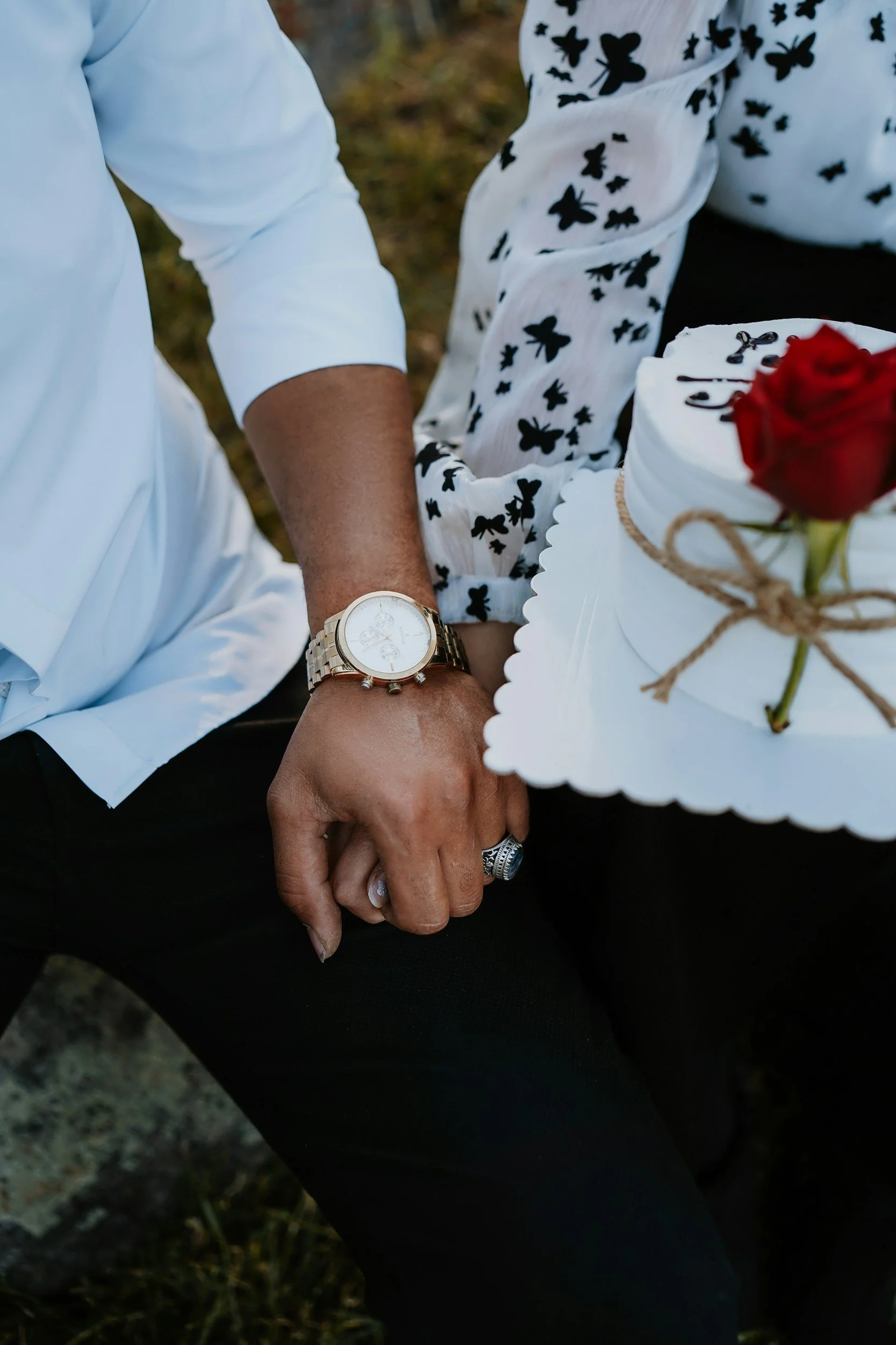 Nahaufnahme eines Paares, das Händchen hält, mit einer Hochzeitstorte und einer Rose im Vordergrund.
