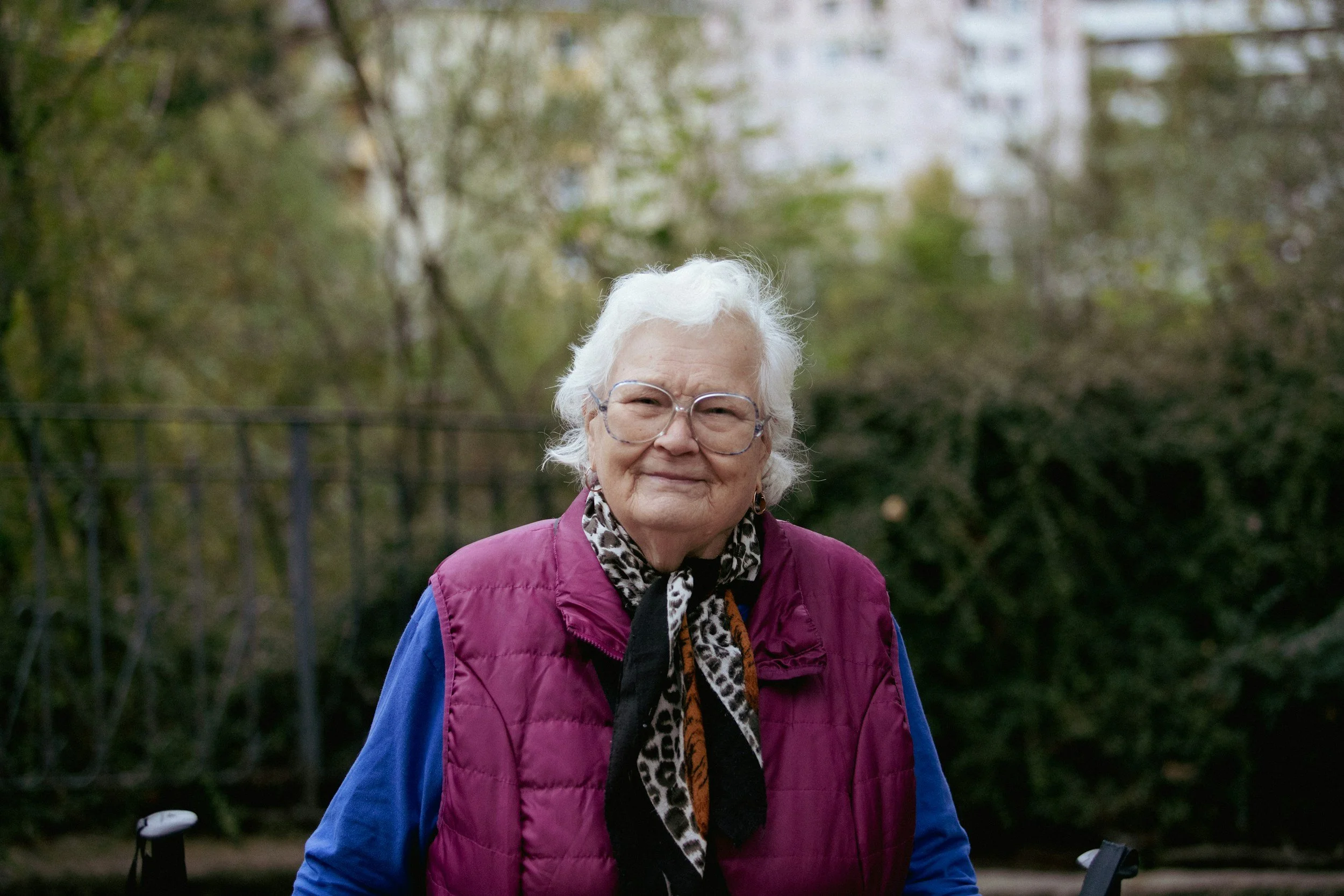 Ältere Frau mit weißen Haaren, Brille und einem Lächeln, trägt eine bunte Jacke mit Leopardenmuster, im Hintergrund sind Bäume und eine Stadt.