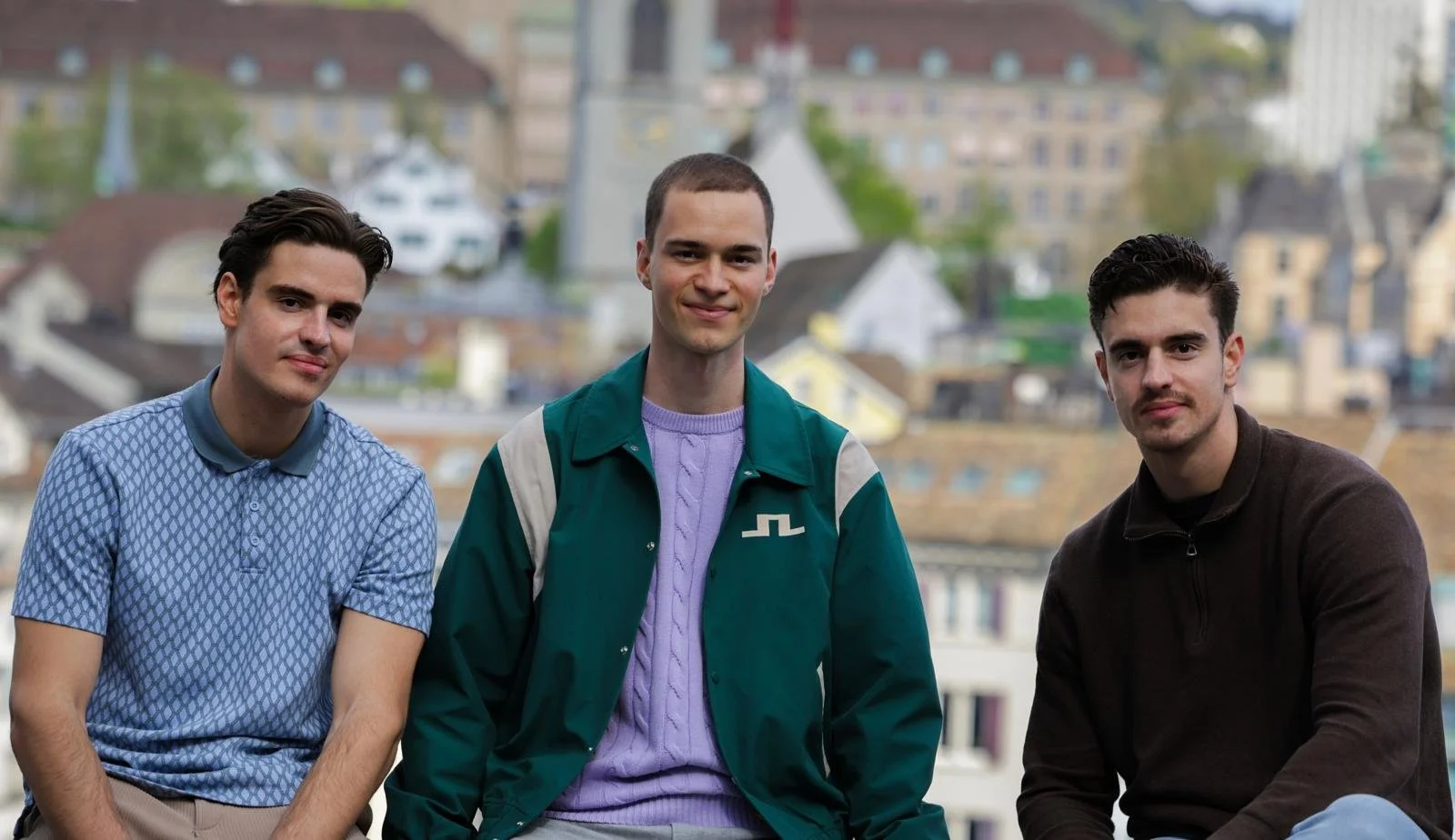 Drei junge Männer sitzen auf einem Balkon mit Stadtblick im Hintergrund.