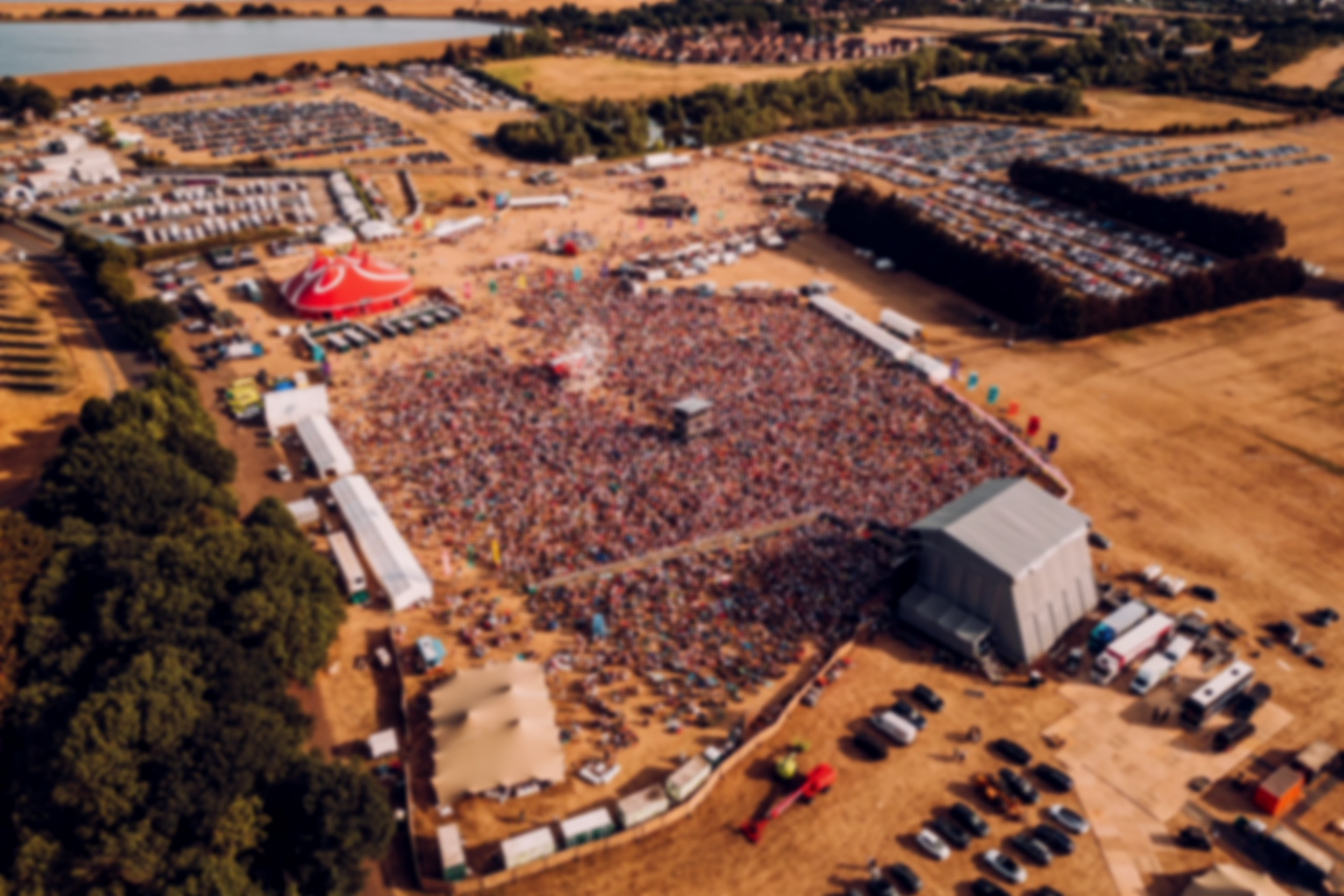 Aerial view of a large outdoor music festival with a crowded stage, tents, and parking lots on a warm day.