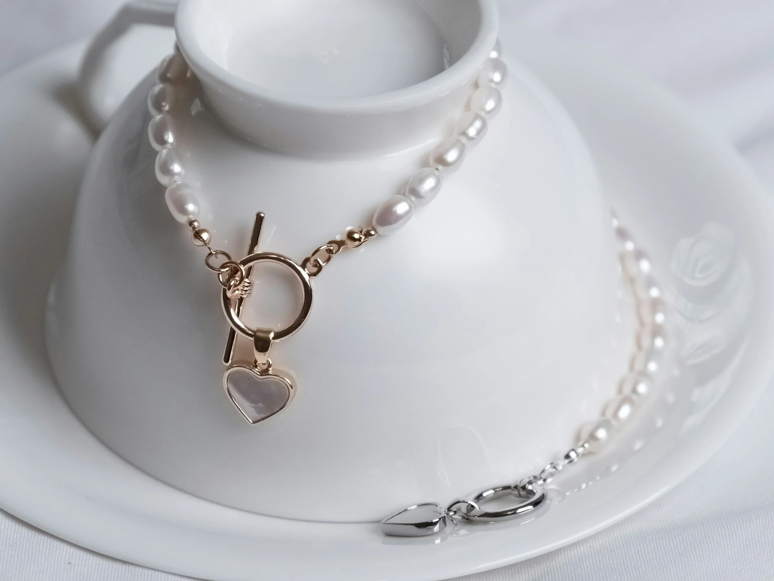 A white teapot with a pearl necklace and bracelet draped over it, on a white plate.
