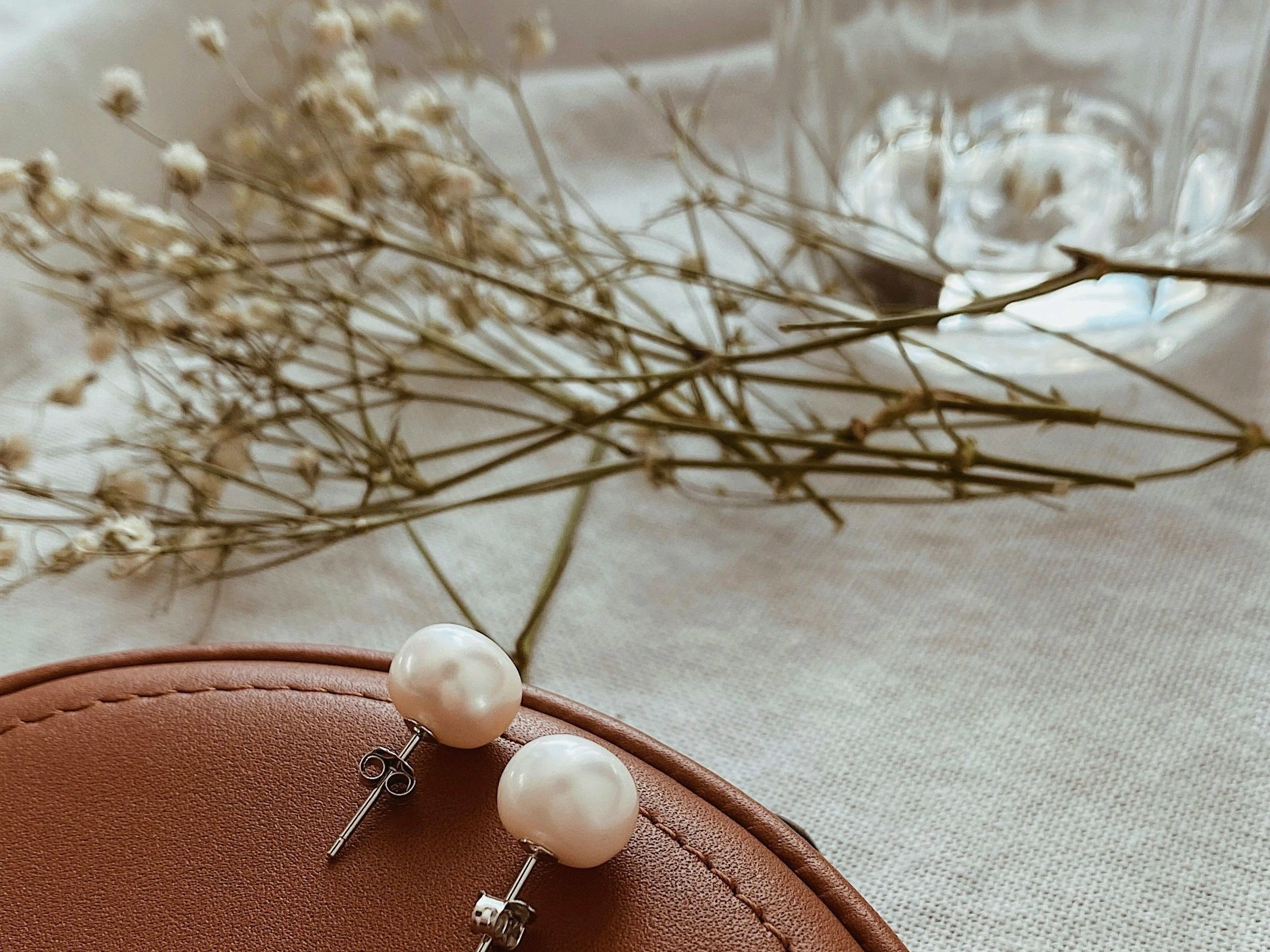 Pearl earrings resting on a brown leather surface with dried flowers and a glass jar in the background.