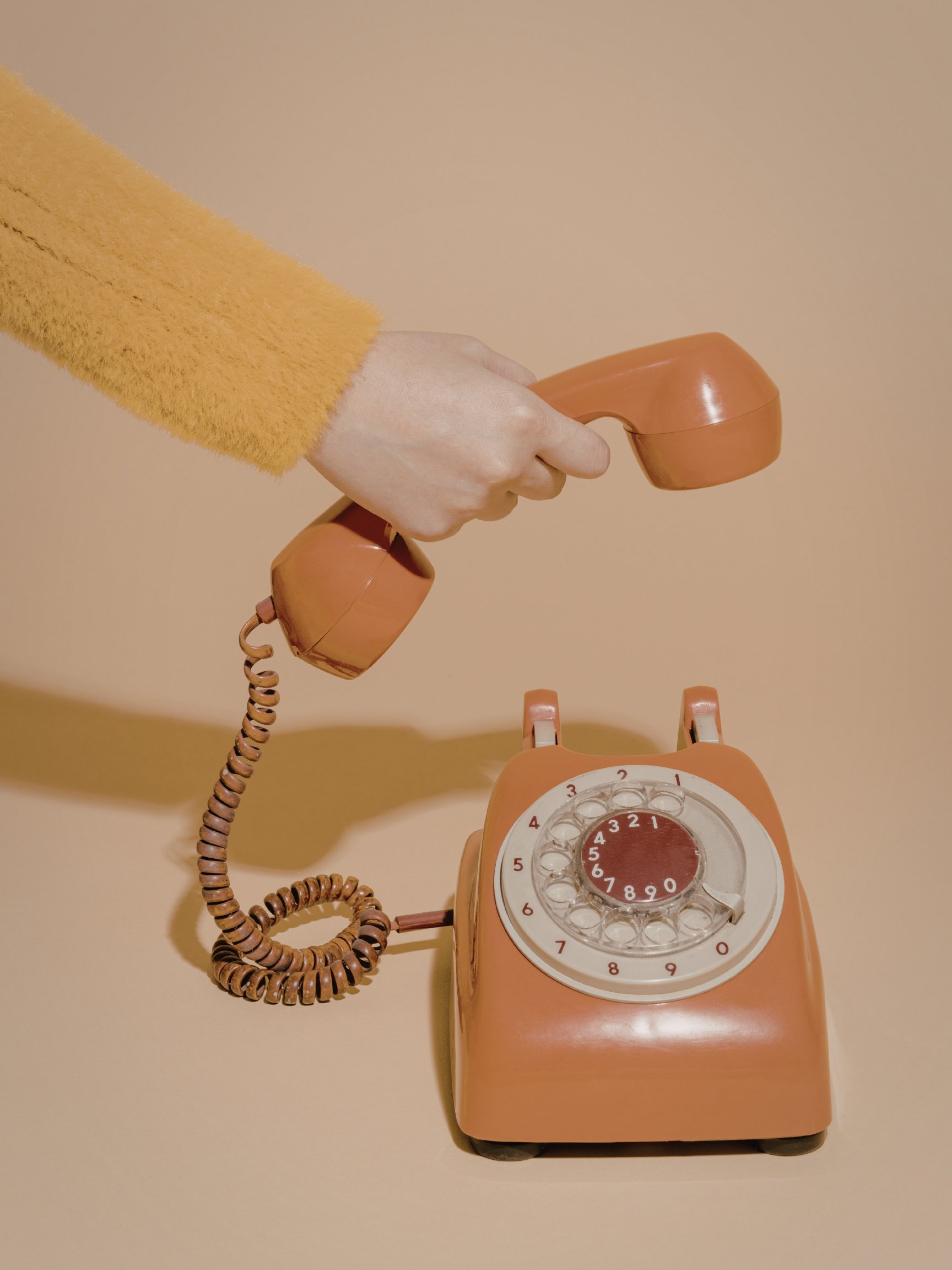 A vintage orange rotary dial telephone with a hand holding the receiver, positioned above the phone.