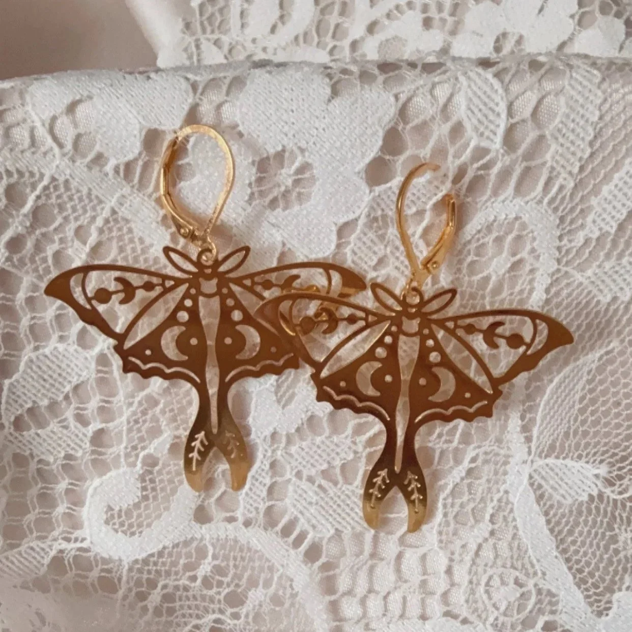 Gold butterfly/ moth-shaped earrings with intricate cut-out designs, resting on white lace fabric.