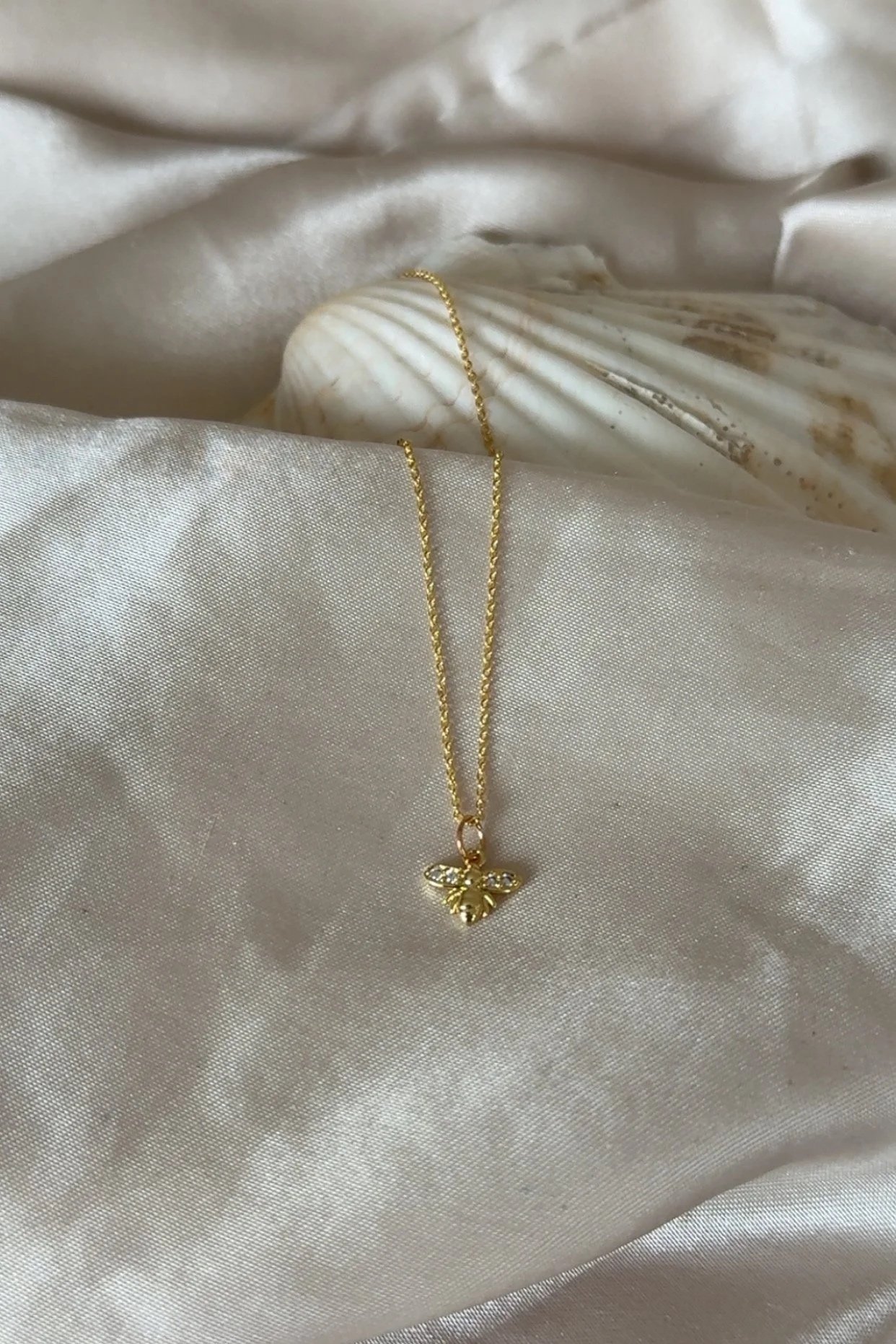 A gold necklace with a bee pendant, resting on a silky beige fabric with a conch shell and white cloth in the background.