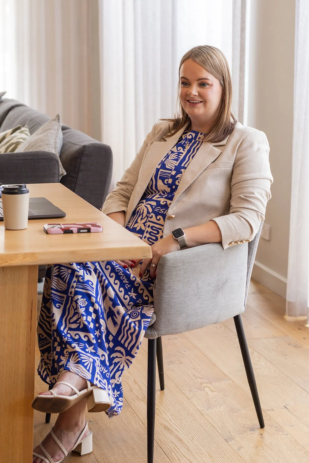 A woman sitting at a wooden table in a modern, well-lit room with beige curtains, wearing a cream-colored blazer over a blue patterned dress, white heels, and a smartwatch.