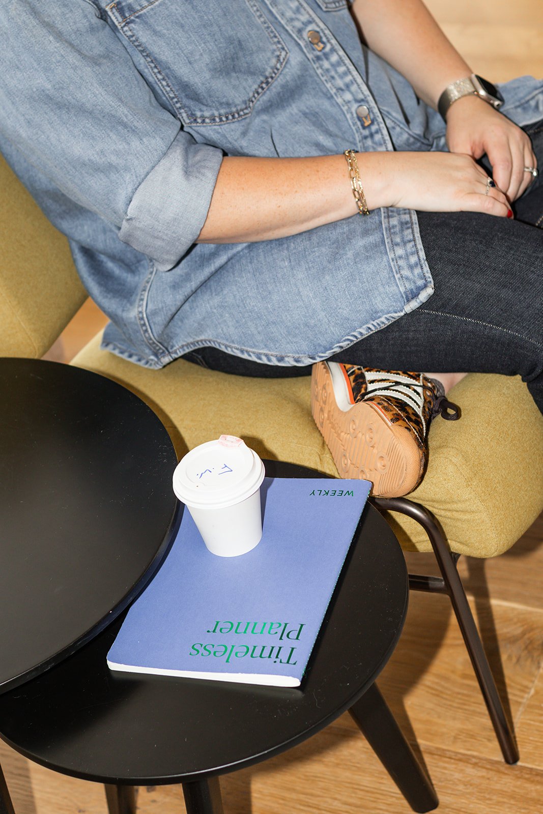 A woman sitting on a yellow couch, wearing a denim shirt, black jeans, and patterned sneakers. On a black round table in front of her, there is a white disposable coffee cup and a blue book titled 'Timeless Plant.'