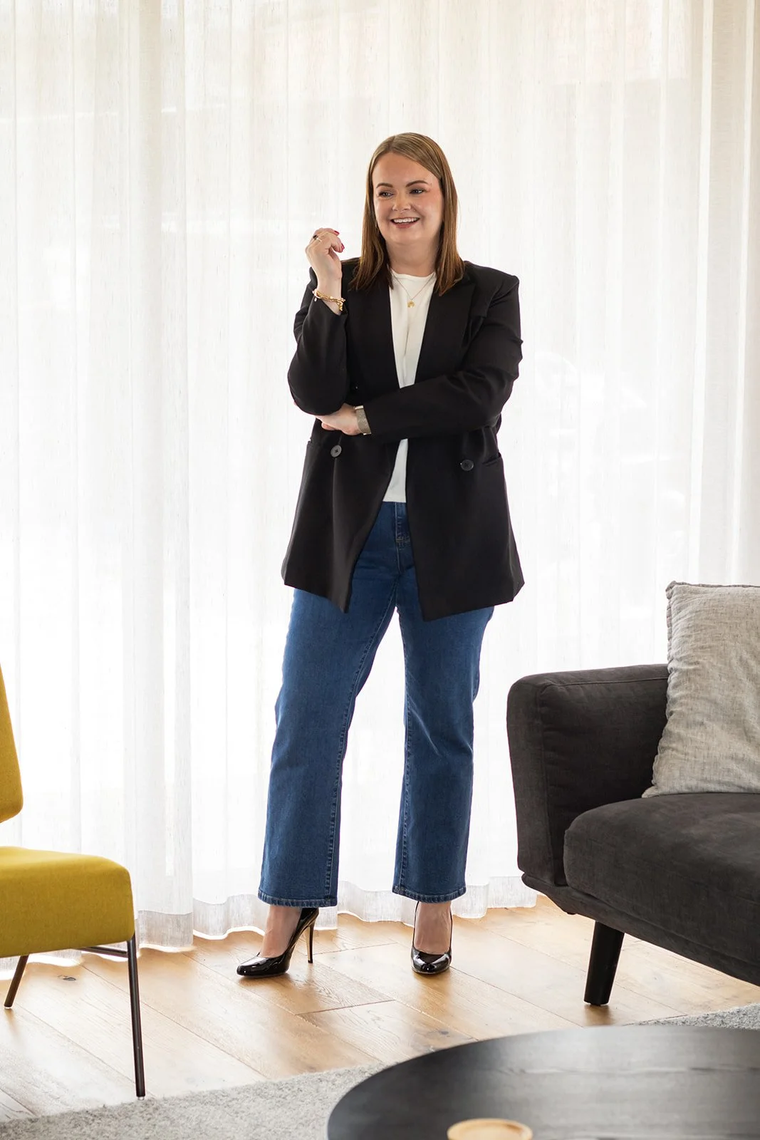 A woman in a black blazer, white top, blue jeans, and black high heels standing in a brightly lit living room with curtains and modern furniture.