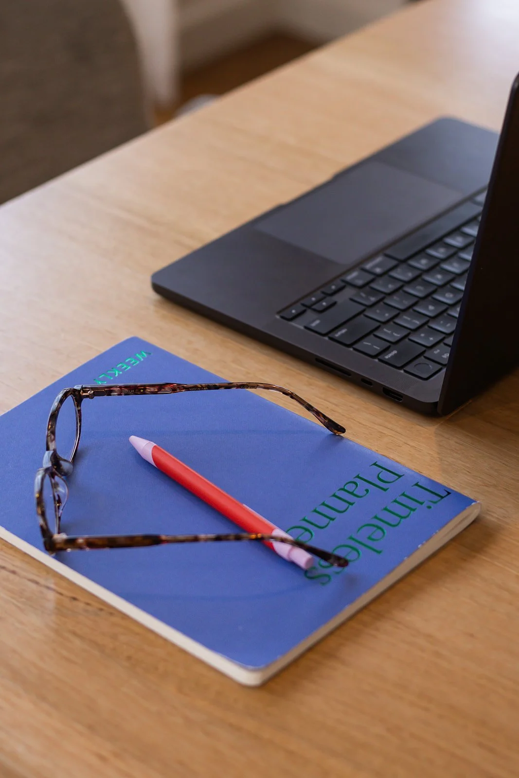 A blue textbook titled 'Taming of the Shrew' on a wooden desk with a red and white pen and tortoiseshell glasses on top, alongside a black laptop.