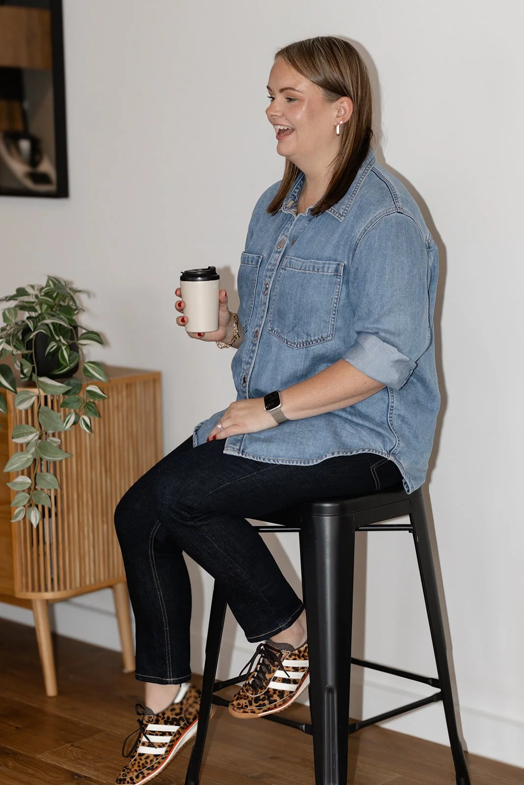 Woman sitting on a black stool, holding a coffee cup, wearing a denim shirt, black jeans, animal print sneakers, and a smartwatch, smiling in a modern indoor setting.