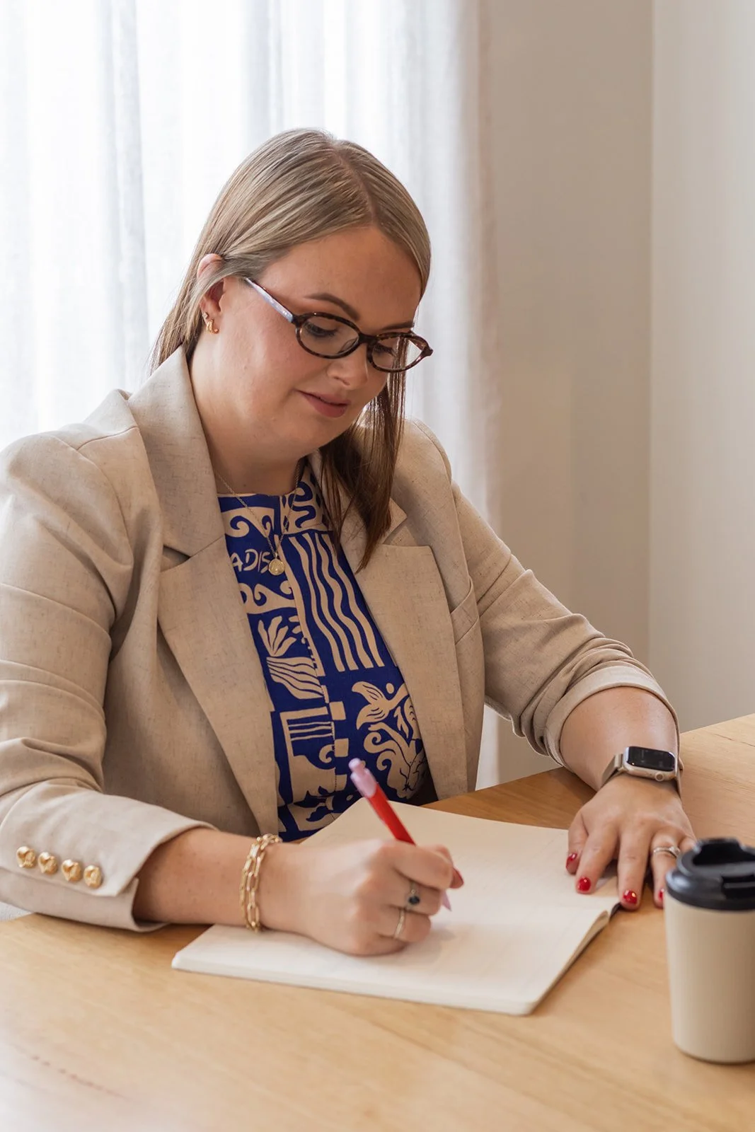 A woman with glasses and blonde hair writing in a notebook at a wooden table, with a disposable coffee cup nearby, in a well-lit room.