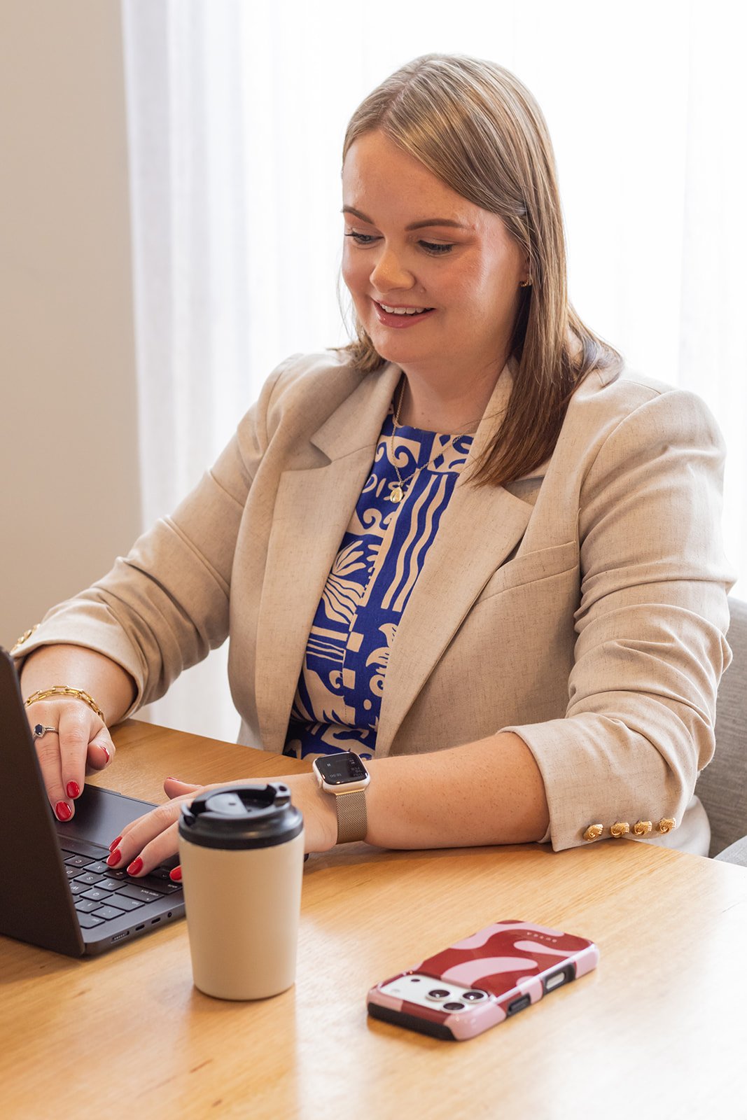 A woman with shoulder-length hair wearing a beige blazer sits at a wooden desk, typing on a laptop, with a coffee cup and a phone on the desk.