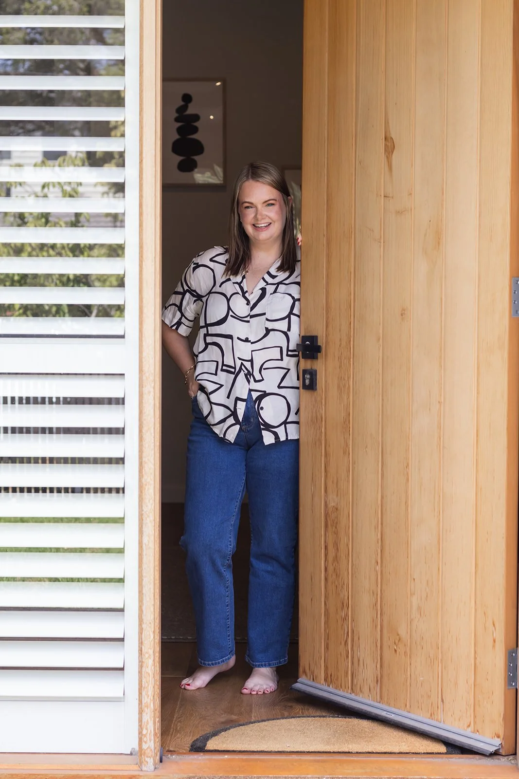 Woman with long brown hair wearing a patterned shirt and jeans standing barefoot at the doorway, smiling.