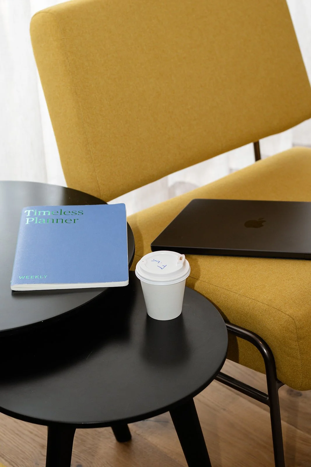 A black round side table with a blue weekly planner, a disposable coffee cup, and a black closed laptop in front of a mustard yellow armchair.