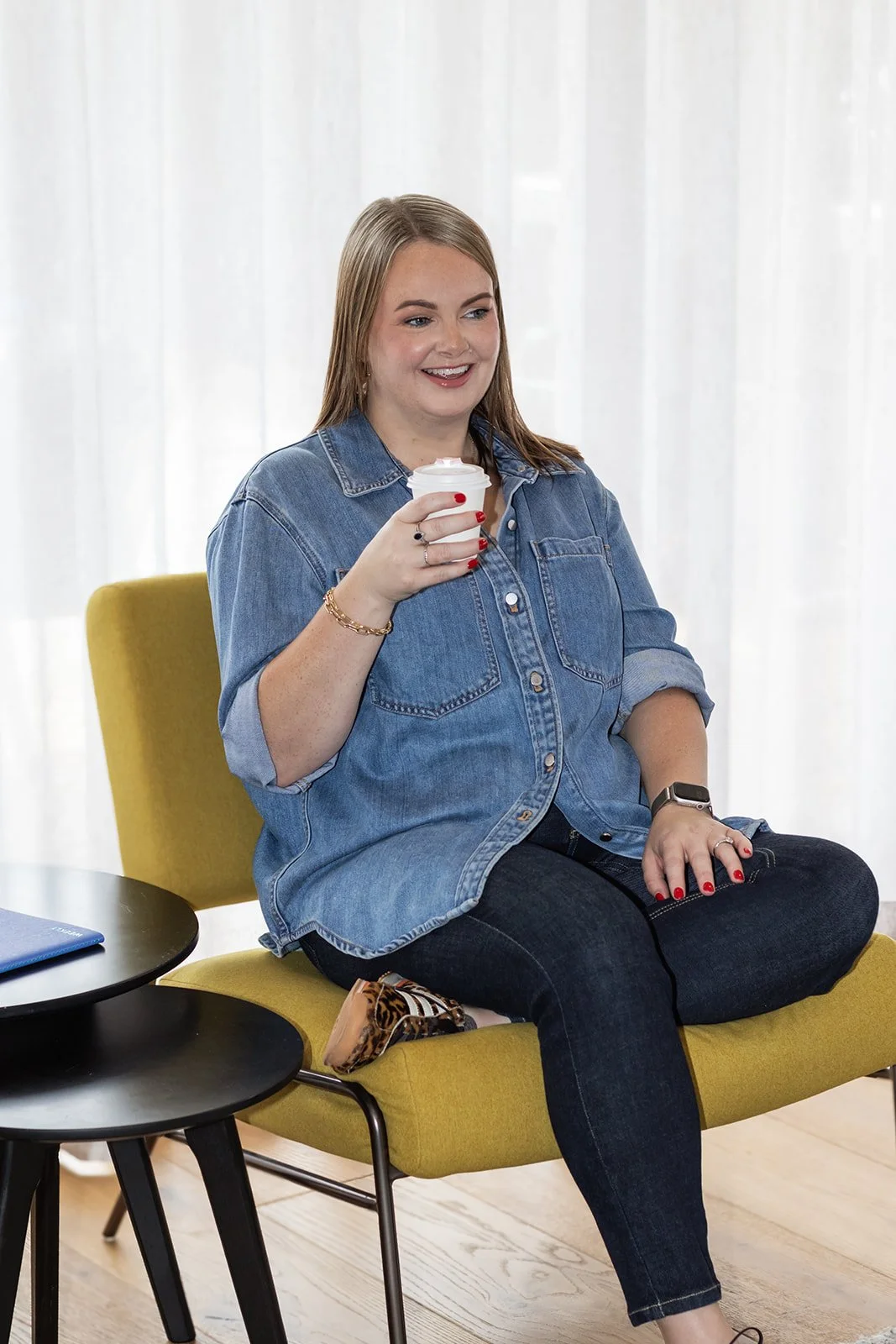 A woman with long blonde hair, wearing a denim shirt and dark jeans, sitting cross-legged on a yellow chair, holding a cup of coffee and smiling.