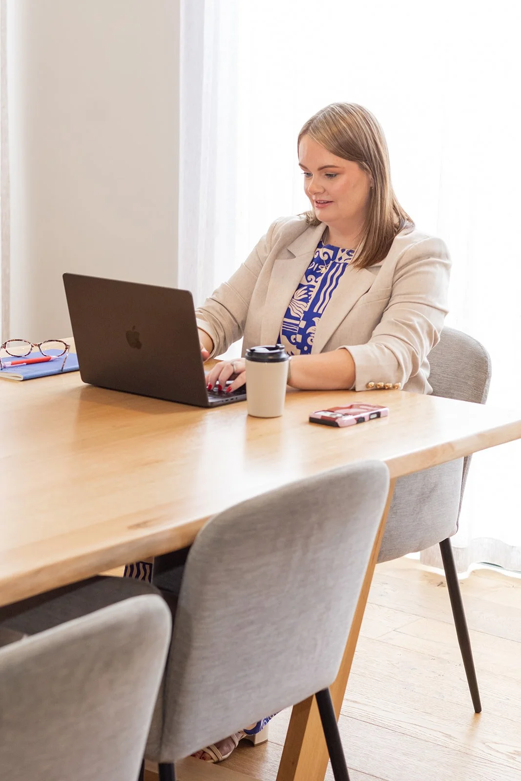 A woman working on a laptop at a wooden table with a coffee cup, smartphone, and eyeglasses nearby in a bright room.