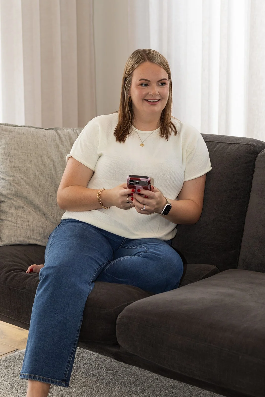 A woman with shoulder-length light brown hair, wearing a white short-sleeved top and blue jeans, is sitting on a dark gray sofa and smiling while holding a smartphone.