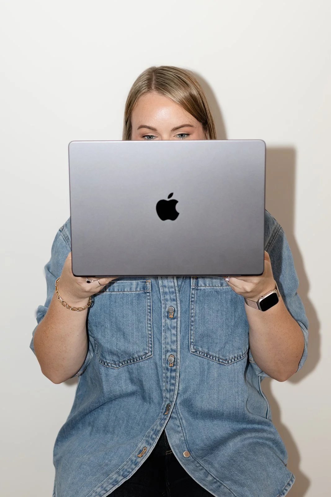 A woman with light brown hair and a denim shirt is looking at a silver MacBook.