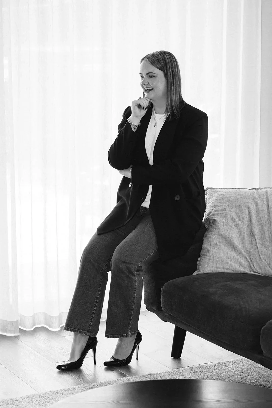A woman sitting on the arm of a sofa, wearing high heels, jeans, and a blazer, smiling and posing with one hand on her chin in a bright room.