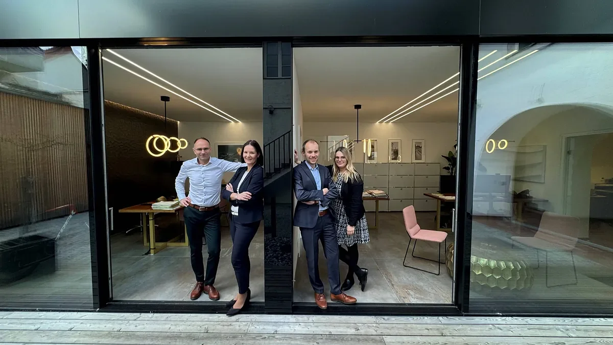 Vier geschäftskleidete Personen stehen vor einem großen Glasausschnitt in einem modernen Bürogebäude, sichtbar sind Tische, Stühle und Büroschubladen im Hintergrund.