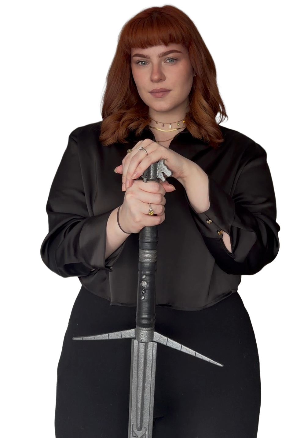A woman with shoulder-length red hair stands confidently holding a sword in front of her. She wears a black satin blouse and layered necklaces.