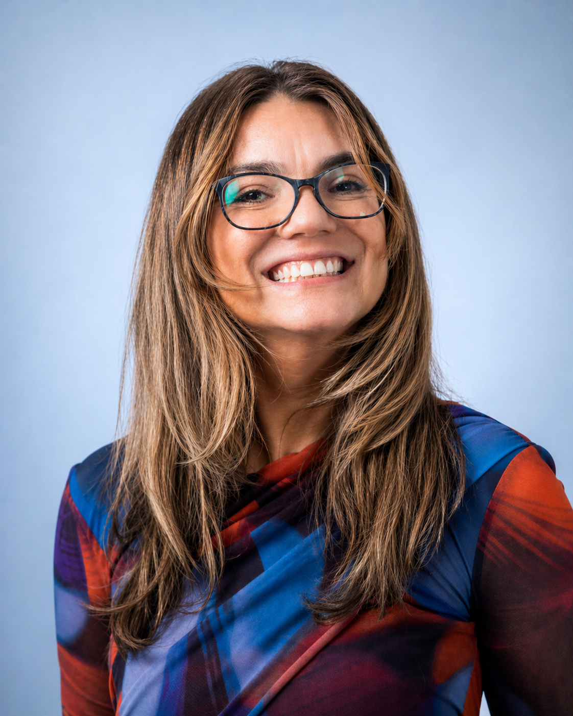 A woman with long, wavy hair and glasses smiling against a light blue background.
