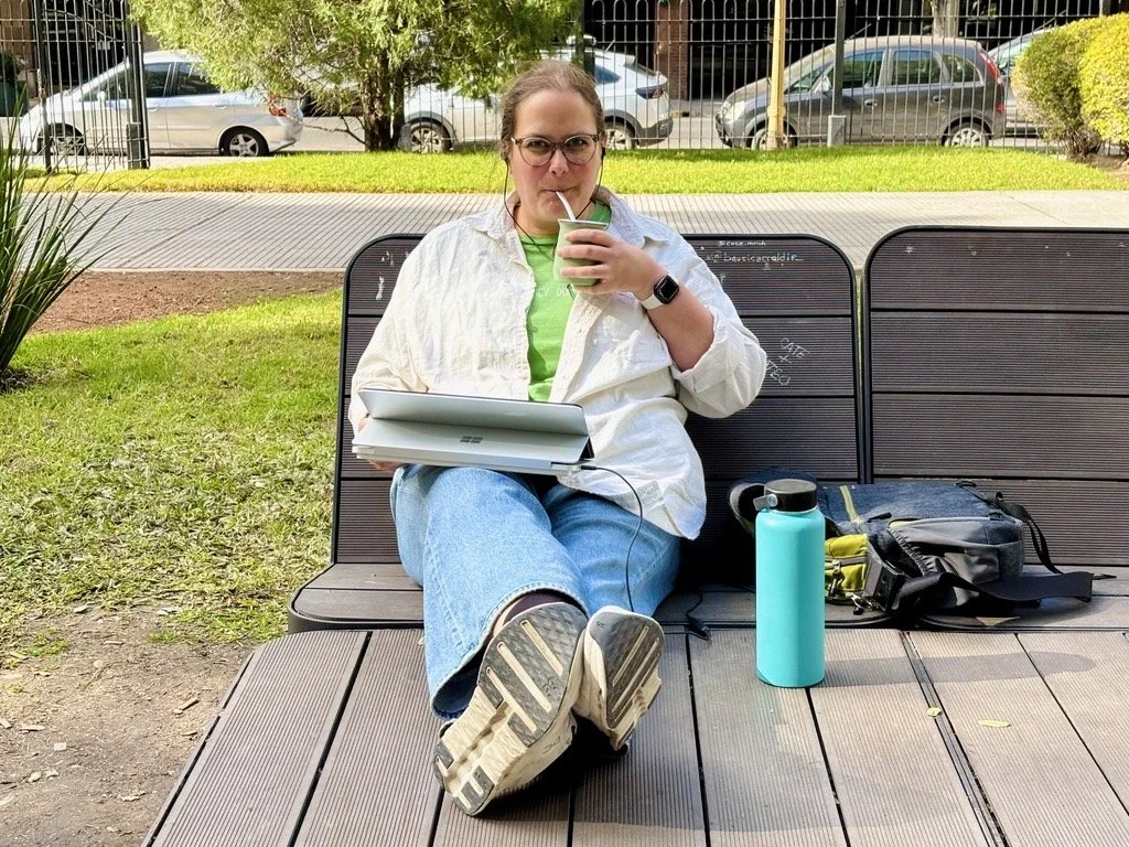 Tamara sitzt auf einer Parkbank in Buenos Aires, die Beine sind ausgetreckt. Auf dem Schoß ein Laptop. Sie trinkt Mate und schaut in die Kamera. Neben ihr stehen eine Wasserflasche und die Laptoptasche.