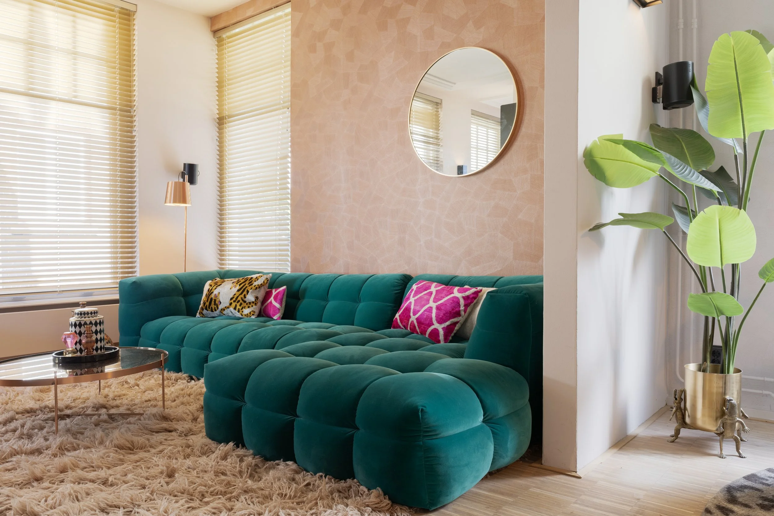 Een woonkamer met grote raampartijen met beige jaloezieën, een groene velvet bank, pinke en dierprint kussens, een ronde zwarte salontafel, een schapenvacht tapijt, een muur met een ronde spiegel, een grote groene plant in een gouden pot en een moderne lamp met koperen accenten.