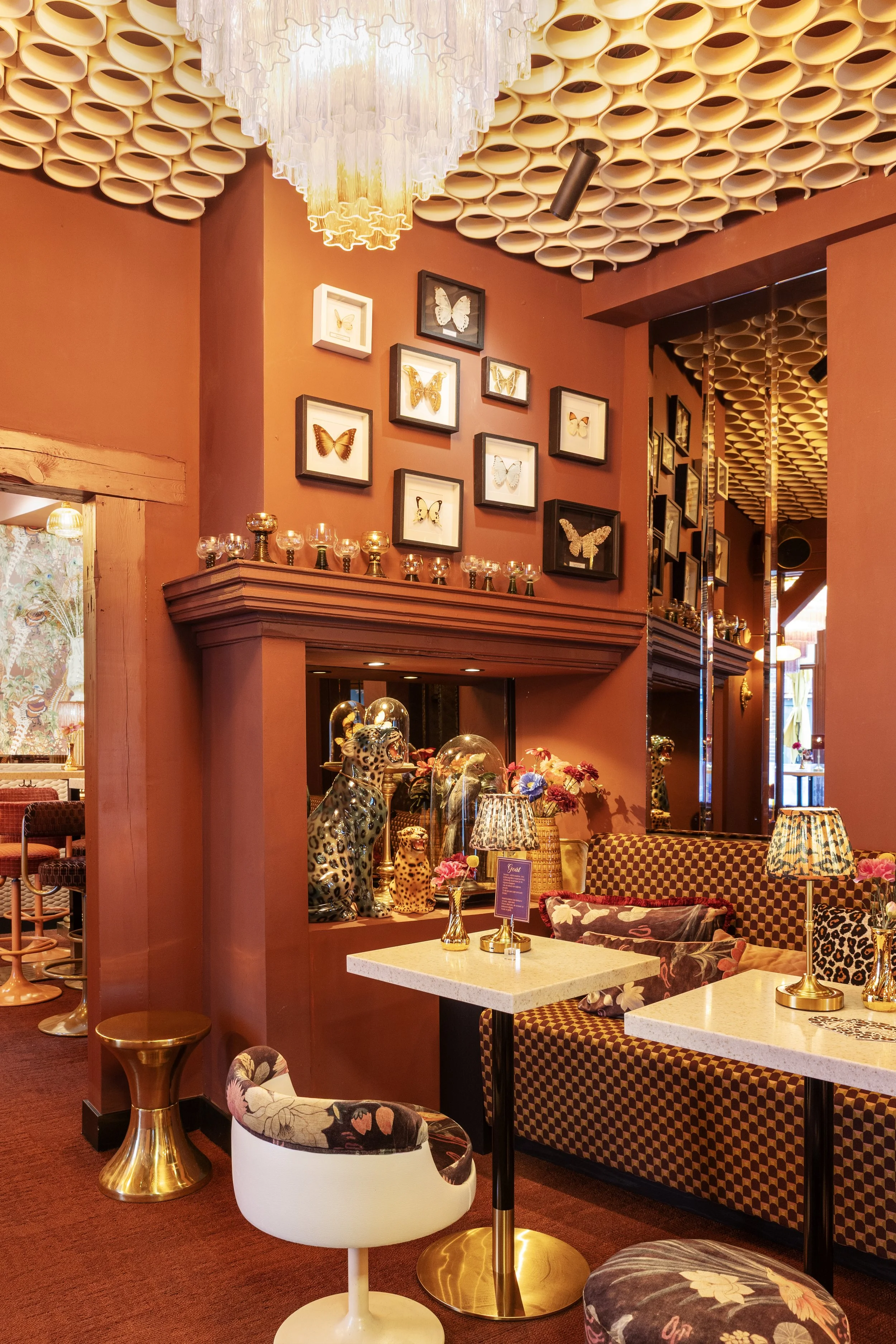 Interieur van een restaurant of café met rode muren, geanimeerde plafonddecoraties van keramische bollen, een grote witte plafondlamp, een muur met ingelijste vlinderillustraties, een verzameling decoratieve beeldjes van een jaguar en andere dieren, en tafels met chaise longue banken en tafellampen.