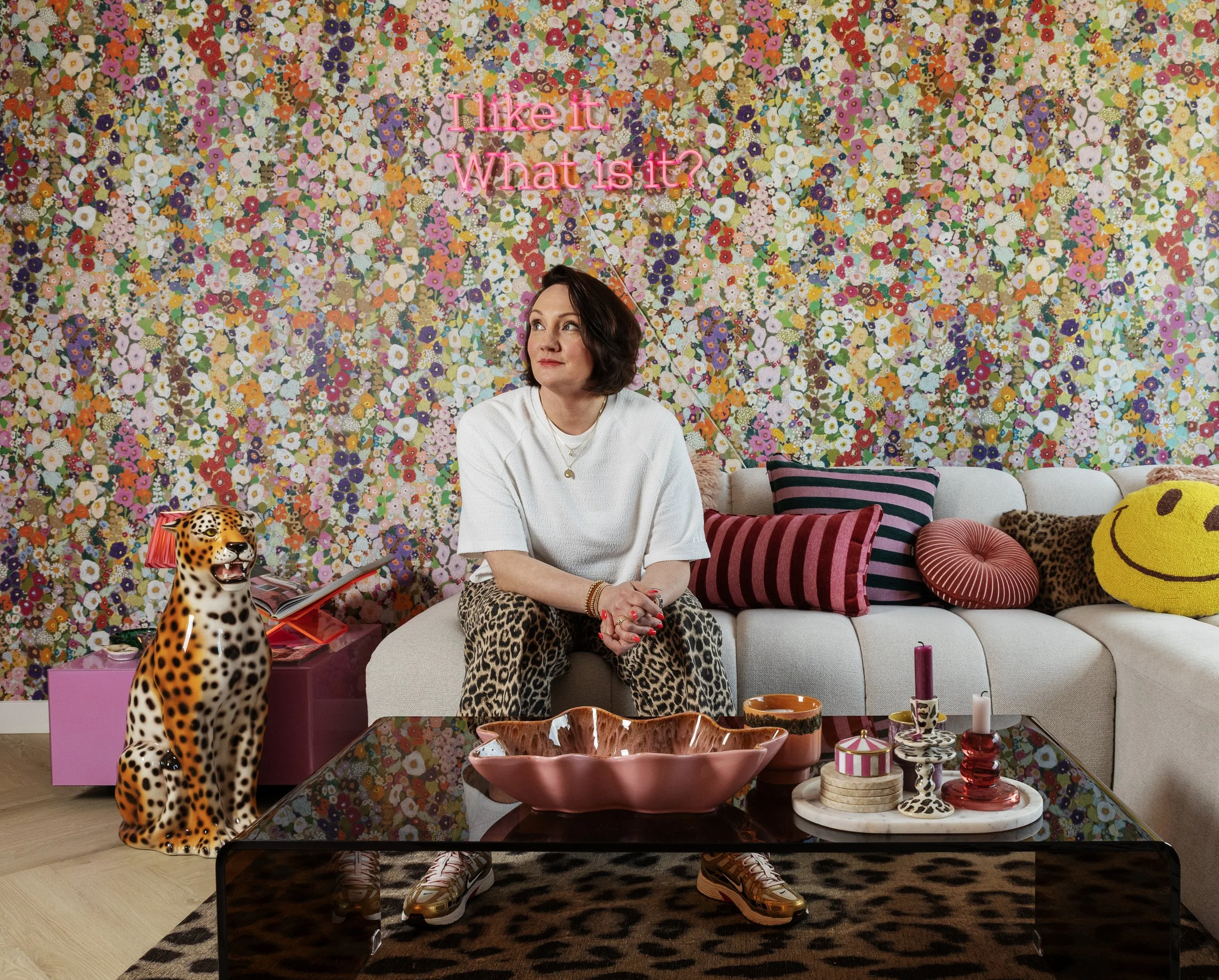 Een vrouw zit op een bank voor een kleurrijke bloemenmuur. Op de tafel voor haar liggen decoratieve voorwerpen, waaronder een leopard-beeld en planten. Op de muur is een neontekst te zien die zegt 'Like it. What is it?'