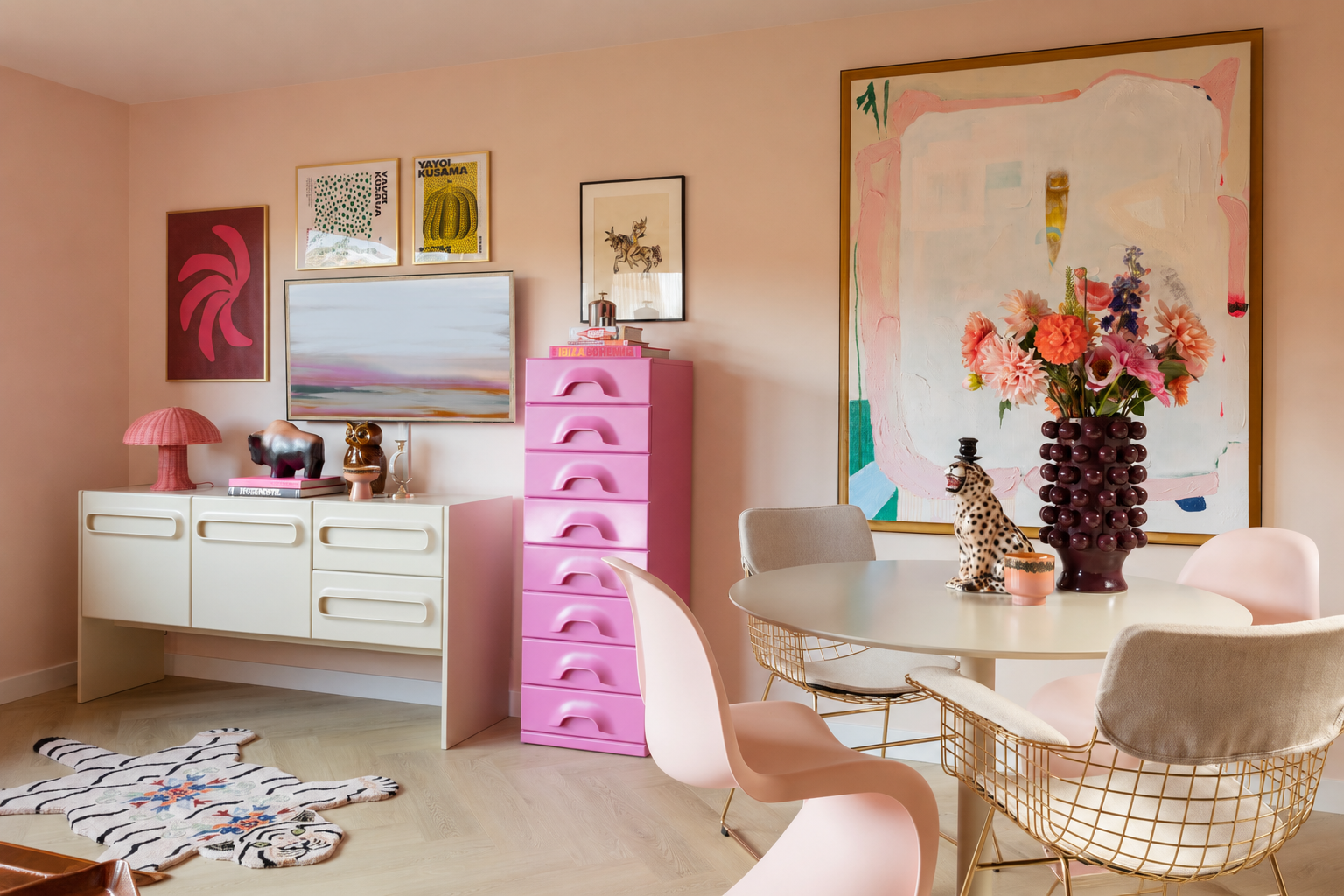 Een woonkamer met een ronde eettafel, diverse gekleurde stoelen, een grote bloemstukken in een vaas, muurkunstwerken, een witte buffetkast met decoratieve items, en een roze ladekast. Op de vloer ligt een schapenvacht uitstalsel.