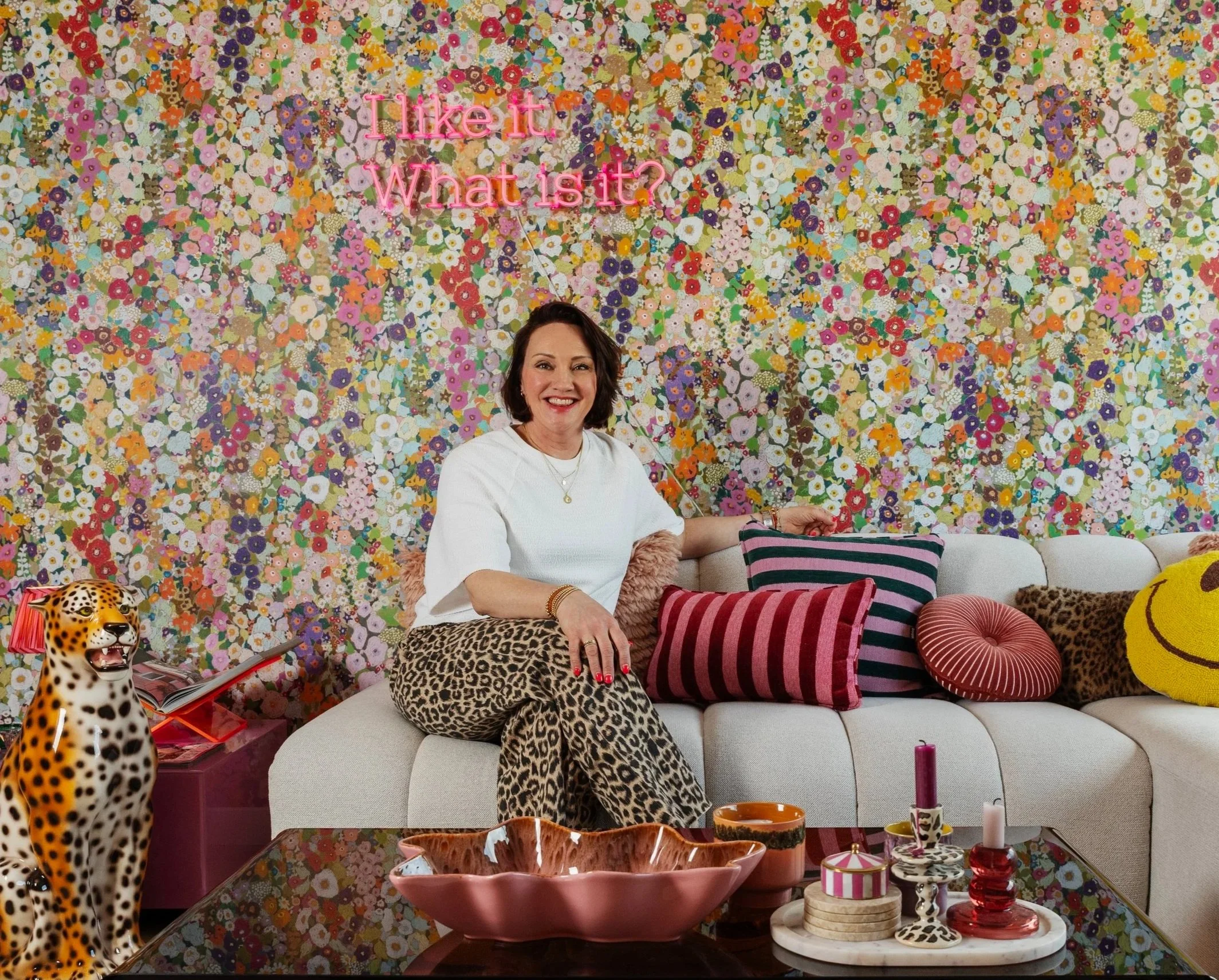Een vrouw die op een bank zit, omringd door kleurrijk behang met bloemen, met decoratieve kussens en accessoires, inclusief een speelgoedluipaard en verfpotten.