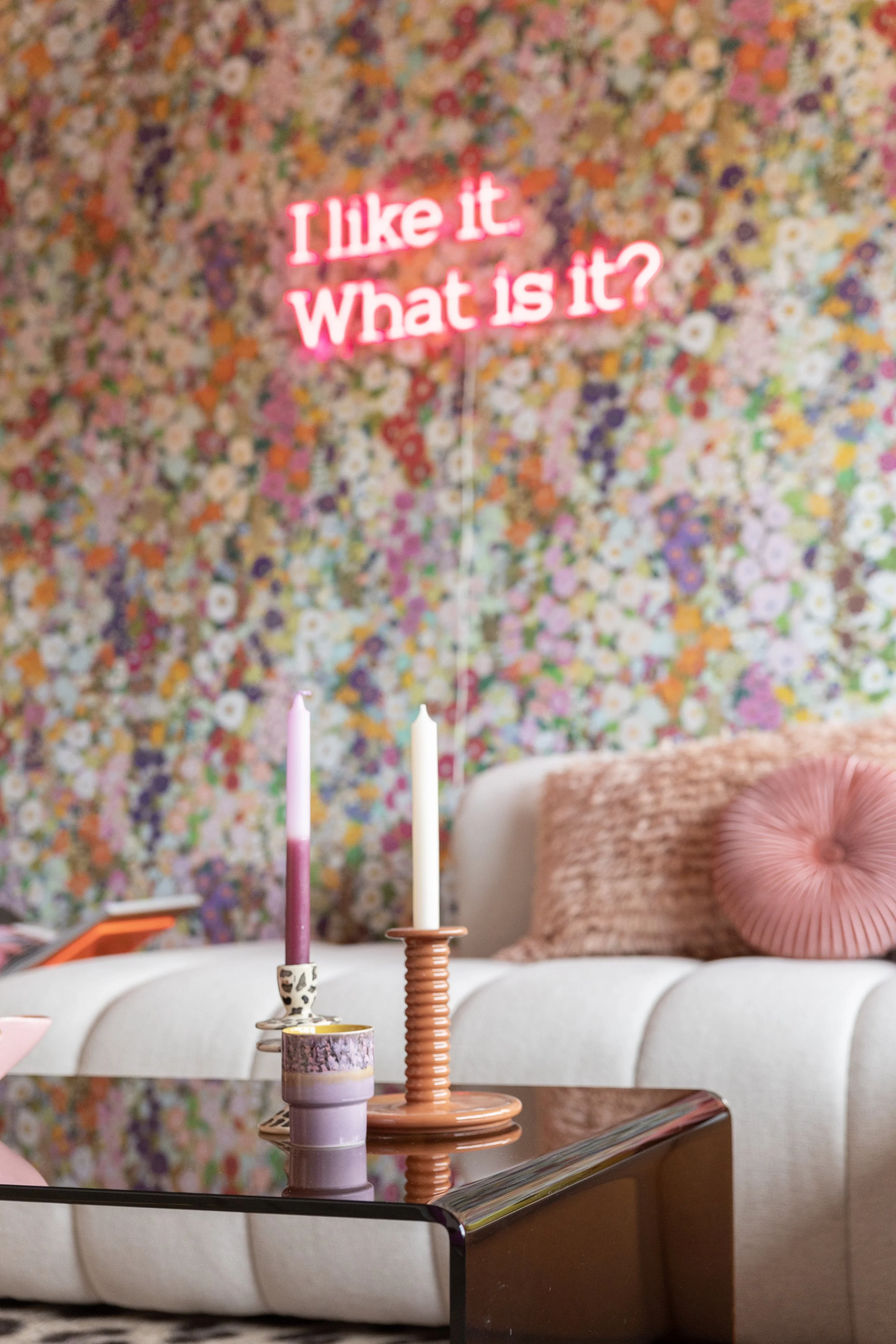 Een woonkamer met een bloemenmuur, een witte bank met een roze kussen en een tafel met verschillende kaarsen. Op de muur hangt een roze neonreclame met de tekst 'I like it, What is it?'.
