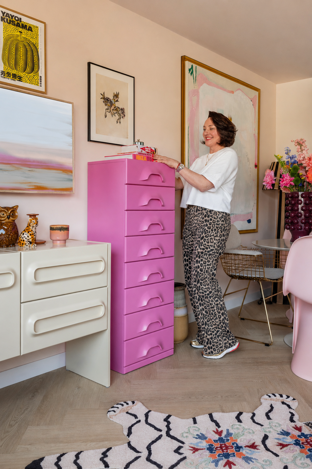 Vrouw die glimlachend staat naast een roze ladekast in een kleurrijk ingerichte woonkamer, omringd door kunstwerken en decoratieve items.