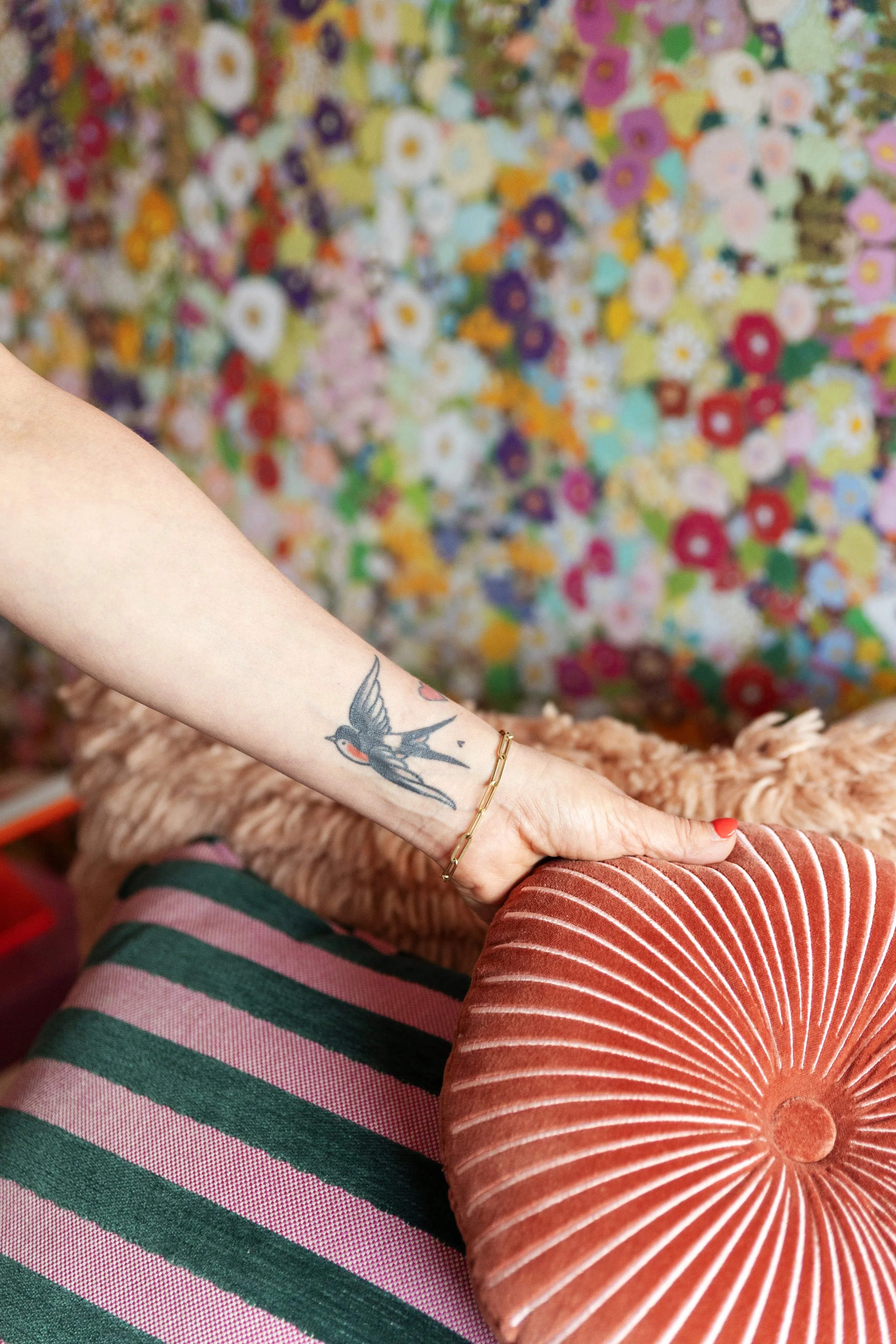 Close-up van een arm met een tattoo van een zwaluw en een armband, rustend op een oranje kussen met rode en witte strepen, met een kleurrijke, bloemrijke achtergrond.