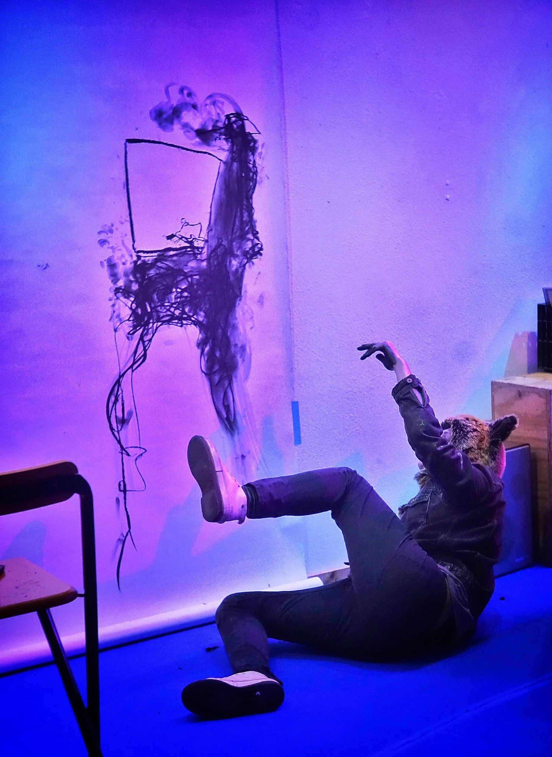 A person with a bear mask is lying on the floor with one leg raised and an arm reaching towards a wall with abstract dragon-like drawing made of black ink. The room has colorful lighting with purple and blue hues.