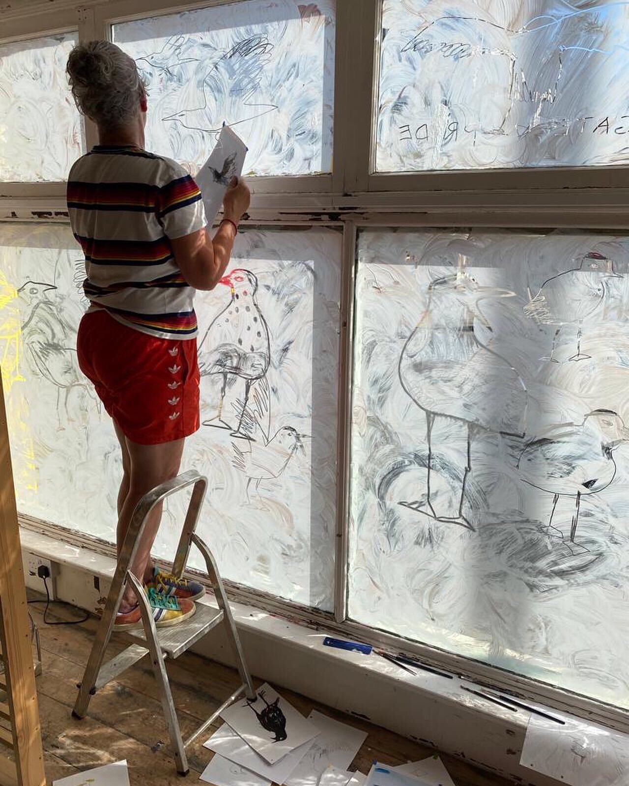 A woman standing on a stepladder, painting large windowpanes with artistic, sketchy drawings of birds, including flamingos and other wading birds, on frosted glass.