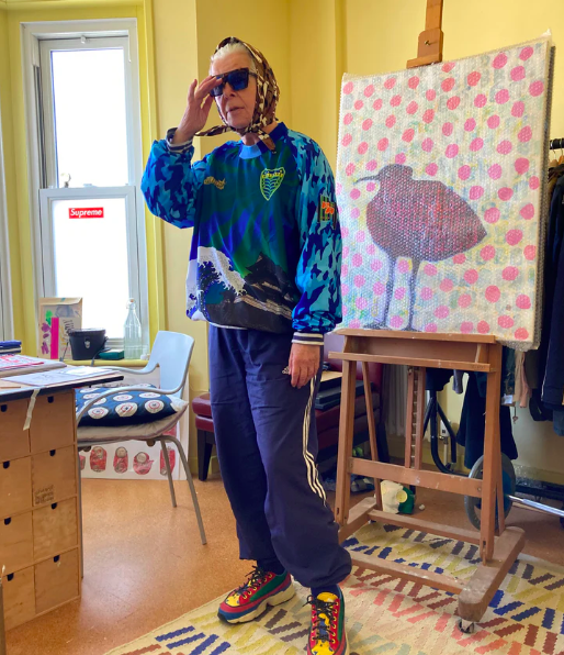 Person wearing sunglasses, a colorful jacket, sweatpants, and sneakers, standing in an art studio with a painting of a duck on an easel nearby.