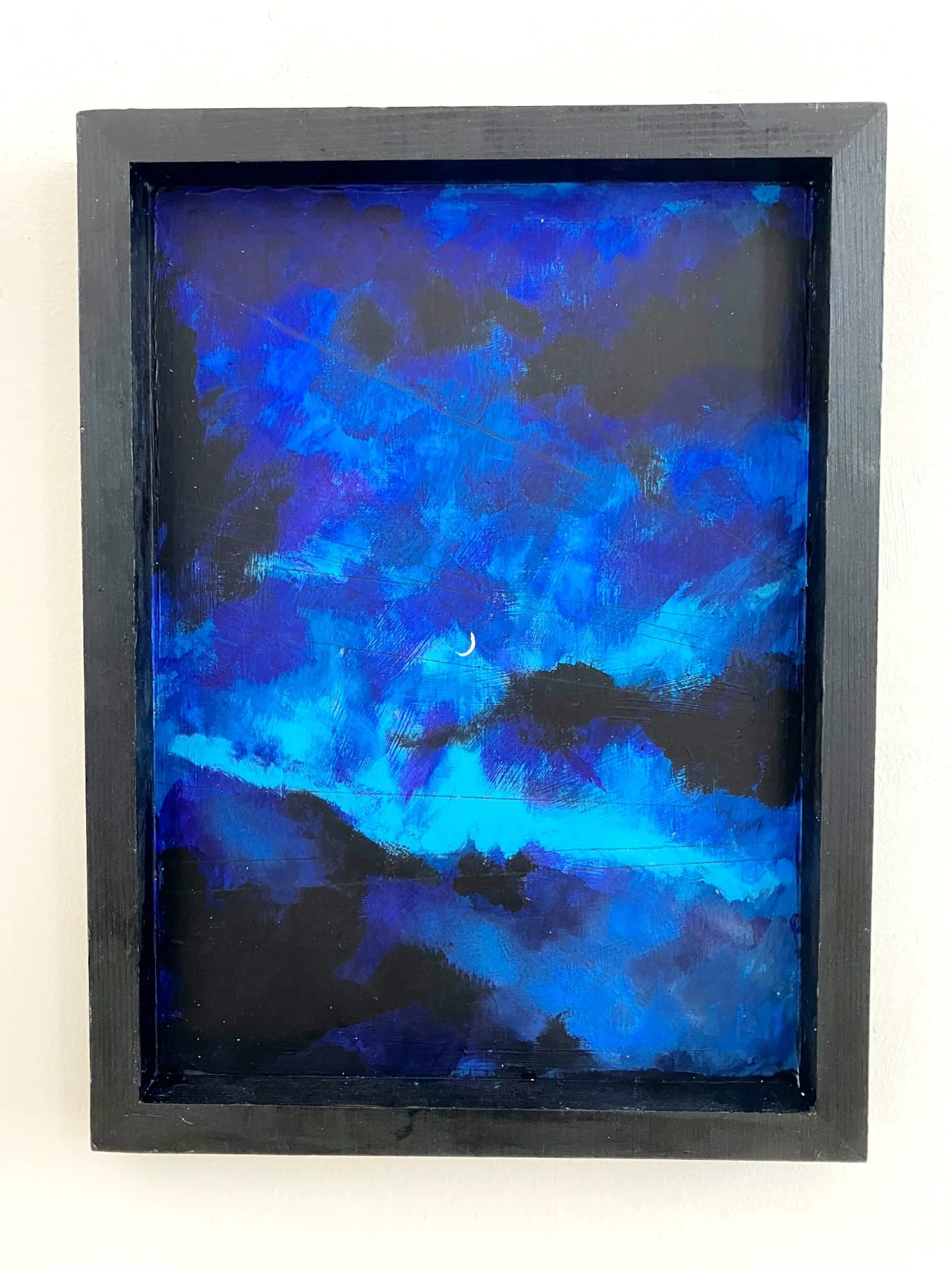 A framed painting of a night sky with dark clouds, a crescent moon, and a blue and black landscape.