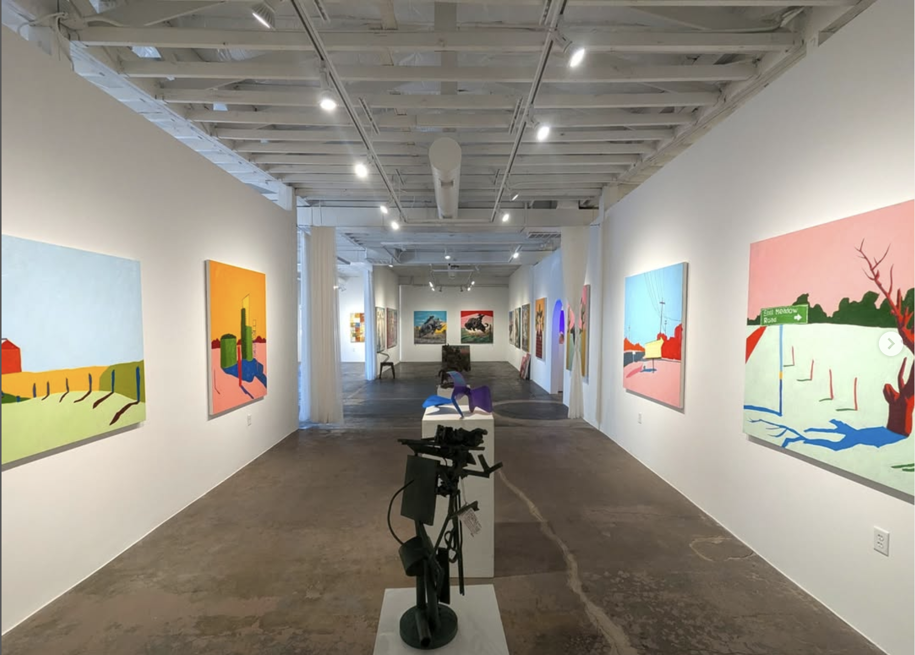 Art gallery with abstract colorful paintings displayed on white walls and sculptures in the center of the room, lit by ceiling spotlights.