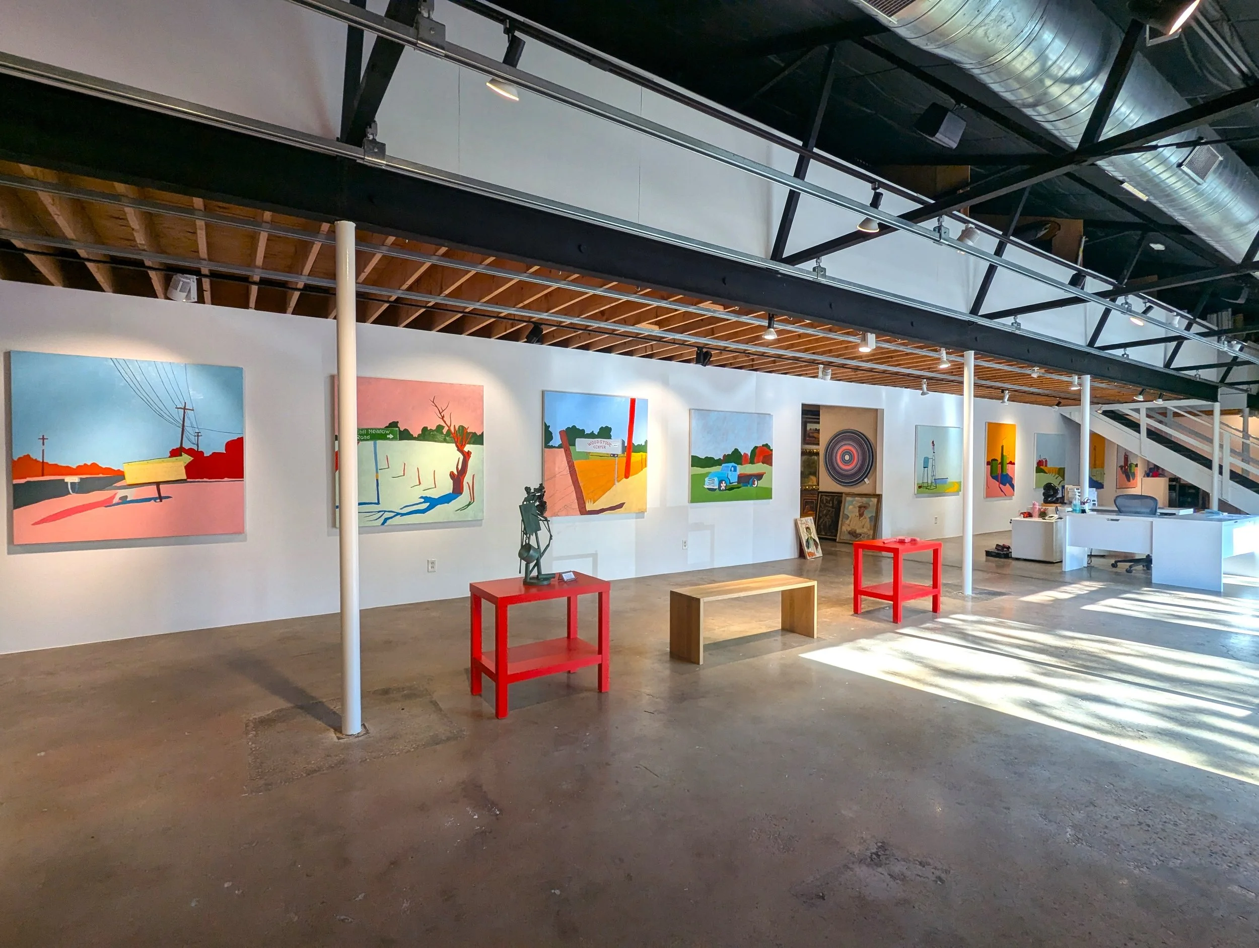 Interior view of an art gallery showcasing colorful landscape paintings on the white wall, with sculptures, benches, and tables in the foreground, and a reception desk in the background under wooden ceiling beams and industrial ductwork.