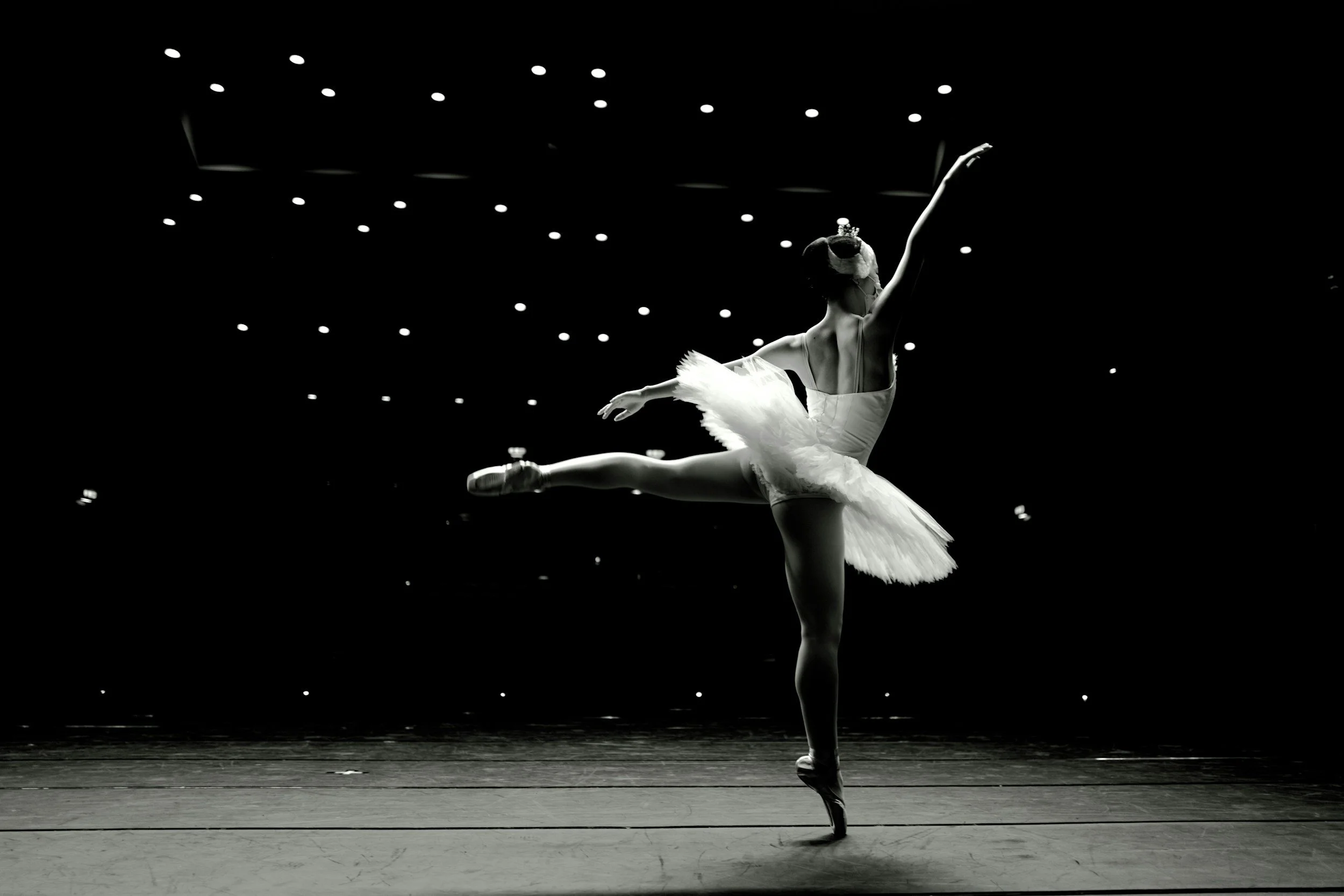 Bailarina en puntas en escenario oscuro con luces en el techo, en posición de baile clásico