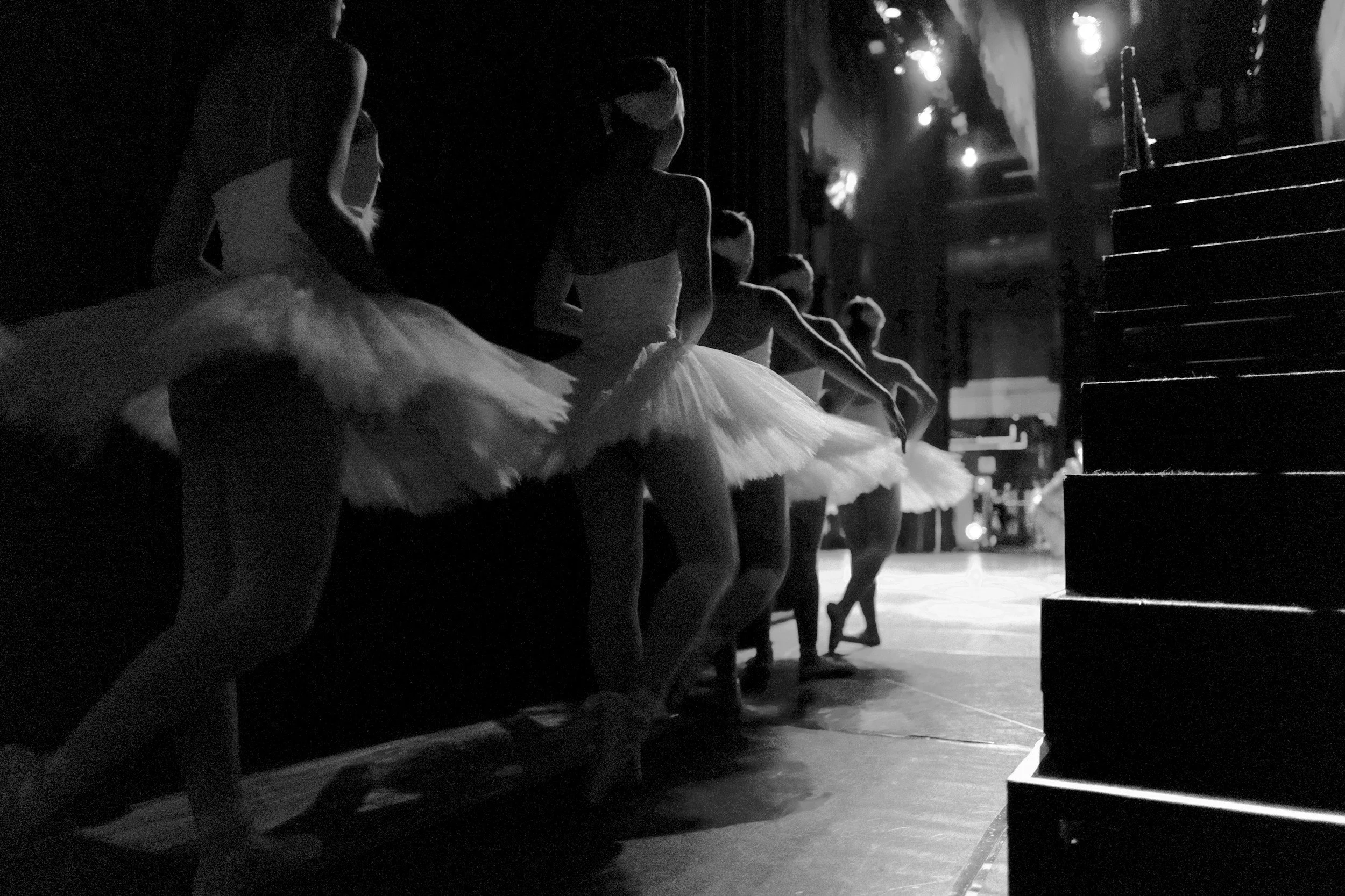 Bailarinas practicando ballet en un escenario con iluminación tenue. Visten tutús blancos y mascarillas negras, alineadas en línea, preparándose para una presentación.