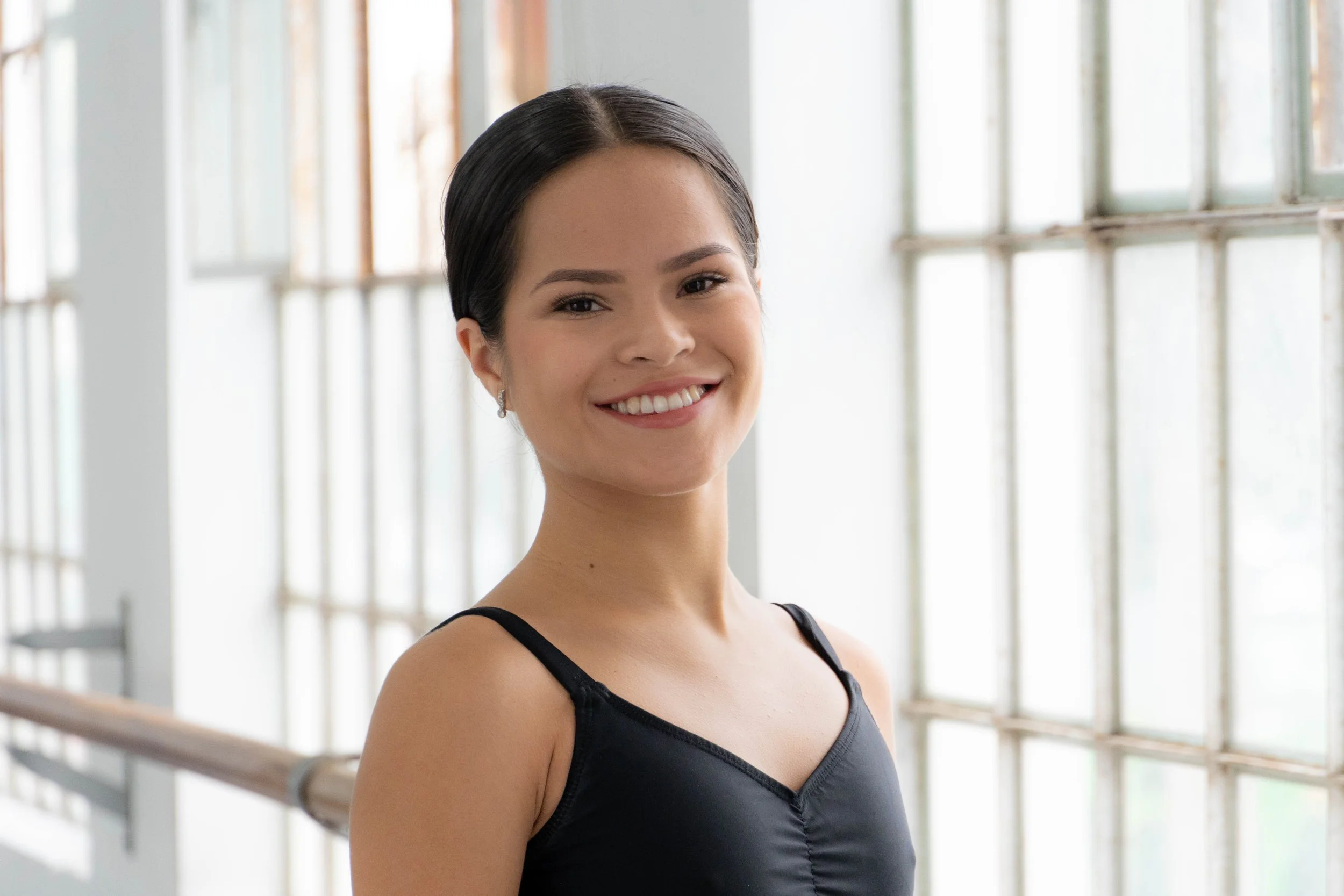 Mujer joven sonriendo de perfil, vestida con un vestido negro, en un espacio con ventanas grandes y barras de metal, posiblemente en un estudio o sala de danza.