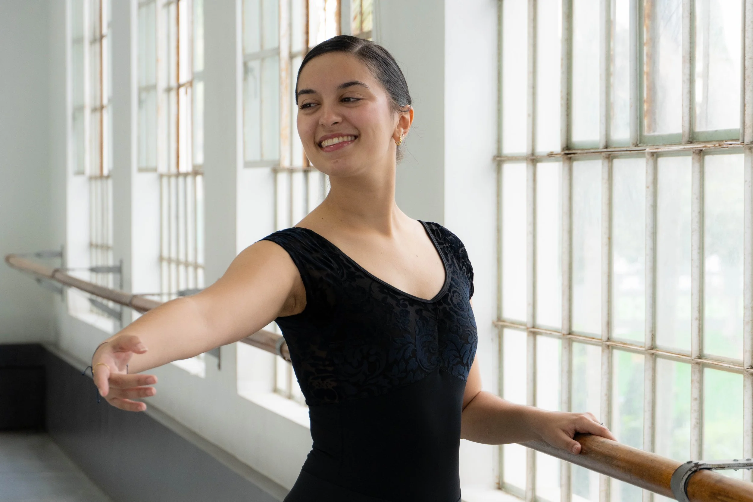 Bailarina sosteniendo una barra en un estudio con ventanas grandes al fondo.