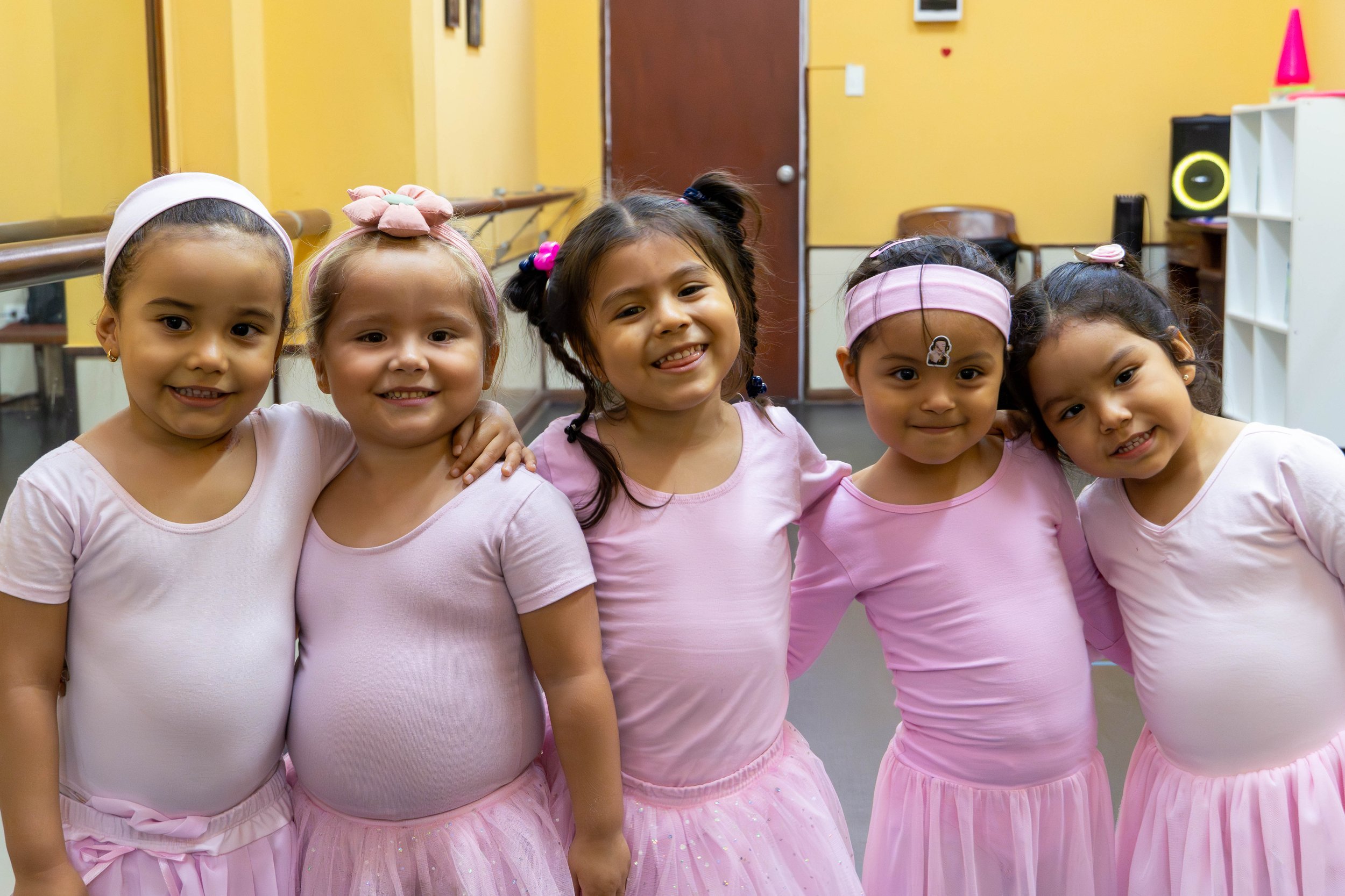 Cinco niñas con peinados y ropa rosa, sonriendo y con los brazos entrelazados en una habitación amarilla.