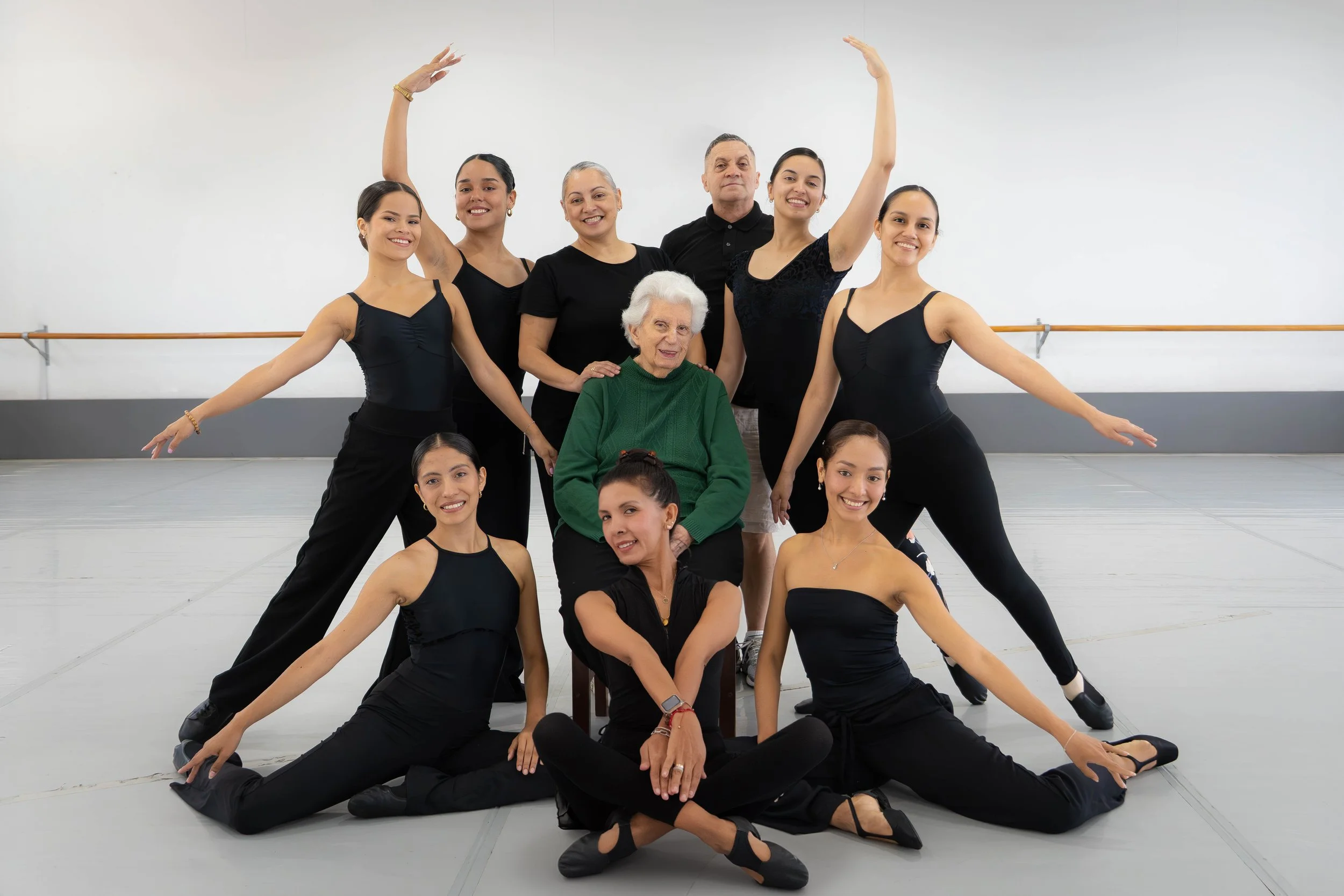 Grupo de personas en un estudio de ballet, con varias bailarinas jóvenes en trajes negros y una mujer mayor con chaqueta verde en el centro, posando con sonrisas y gestos expresivos.