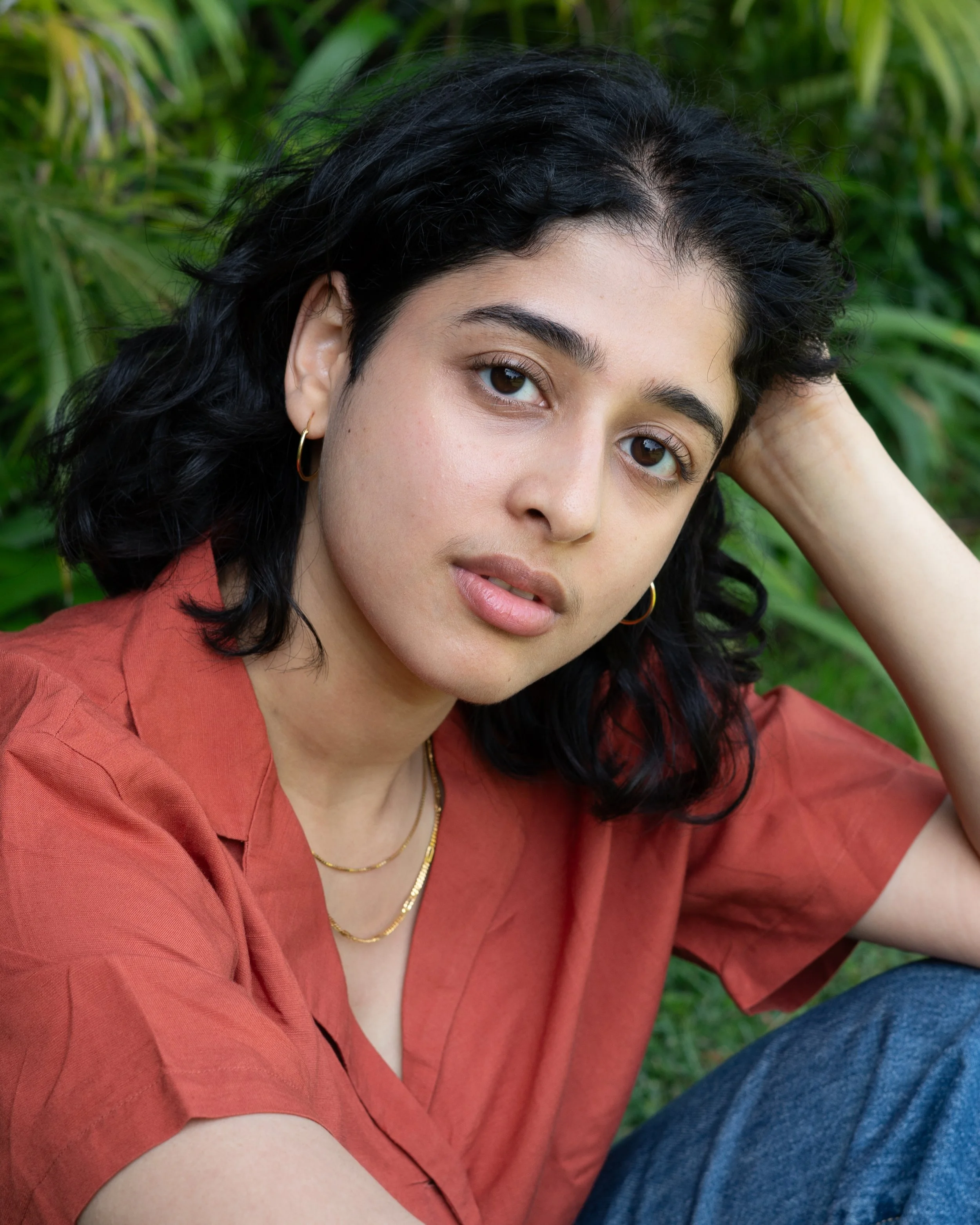 A young woman with black curly hair and hoop earrings, wearing a rust-colored shirt and layered gold necklaces, poses outdoors against a backdrop of greenery.