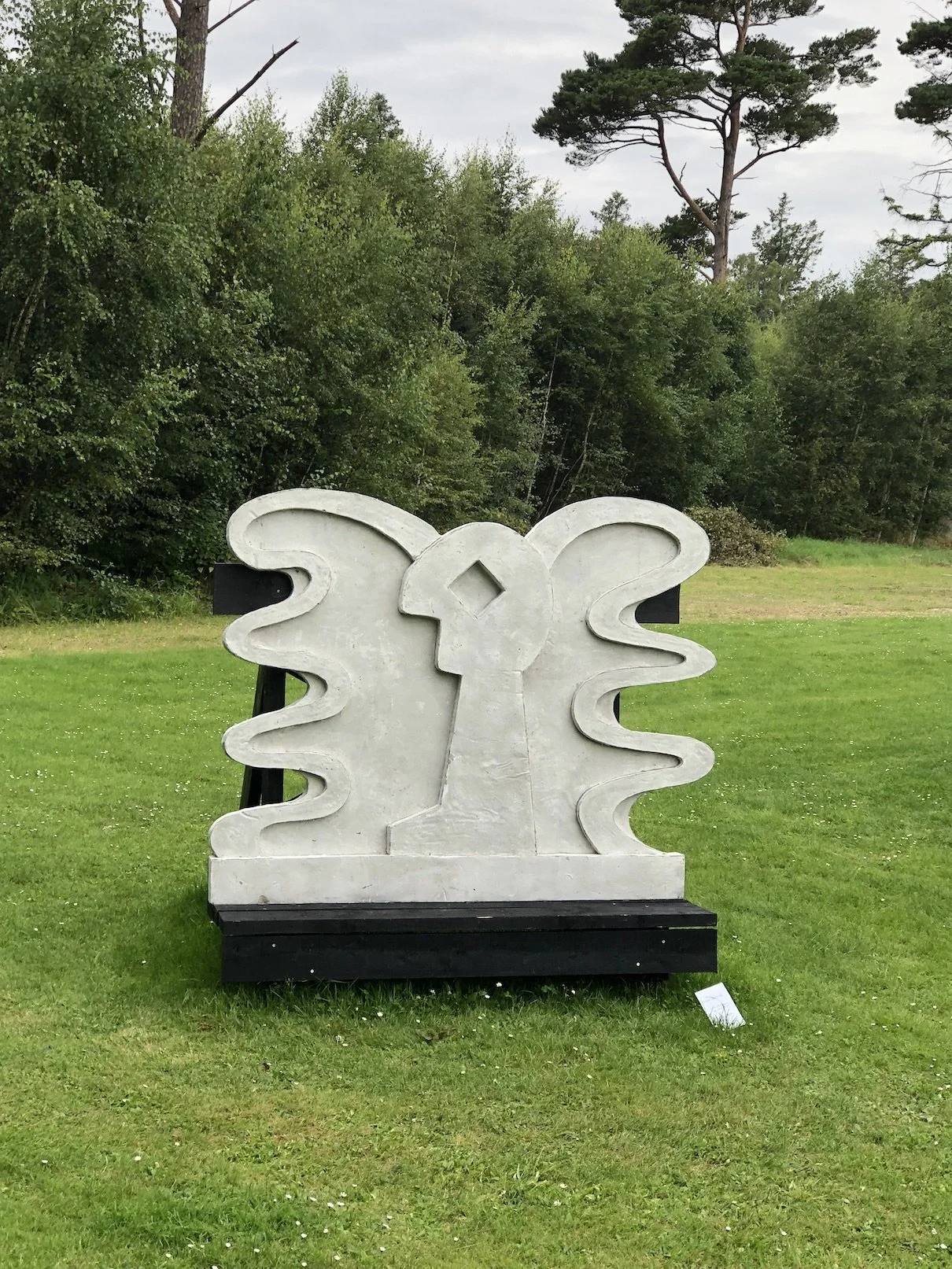 Skulptur med to ansigter, der vender mod hinanden placeret på en græsplæne med træer bagved.