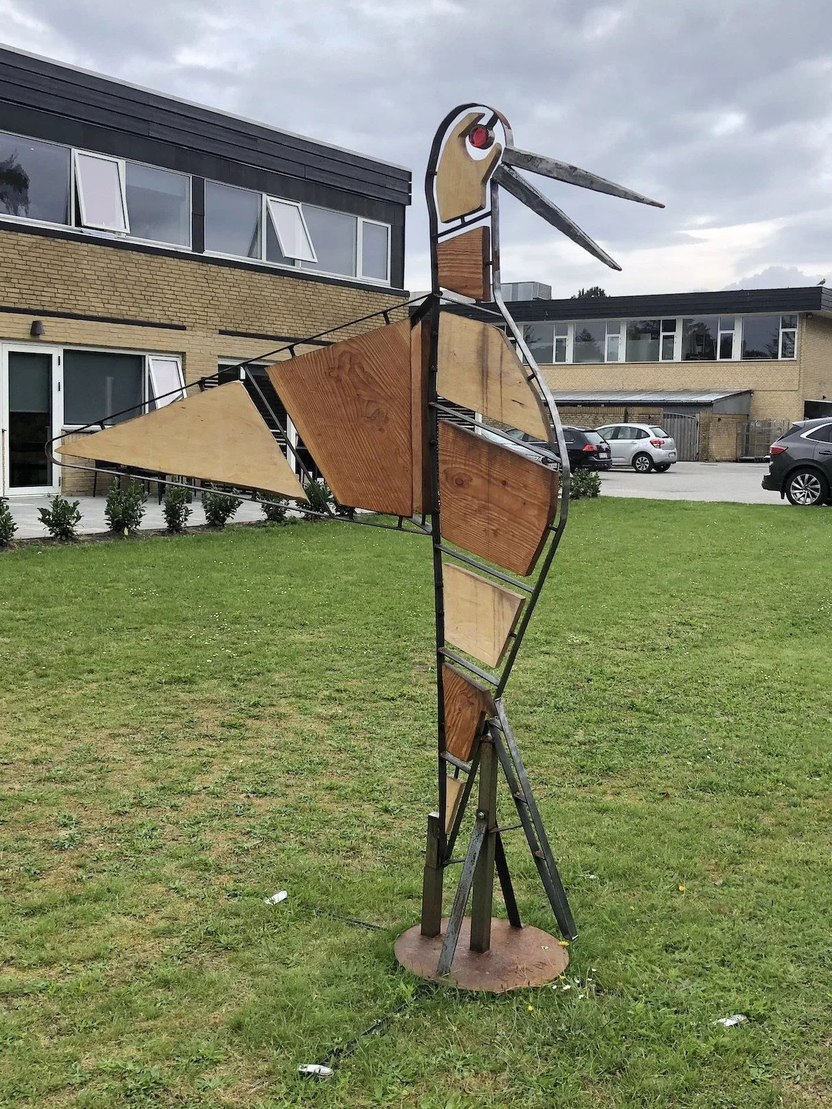 Skulptur af en fugl lavet af metal og træ, placeret på en græsplæne foran moderne bygninger.